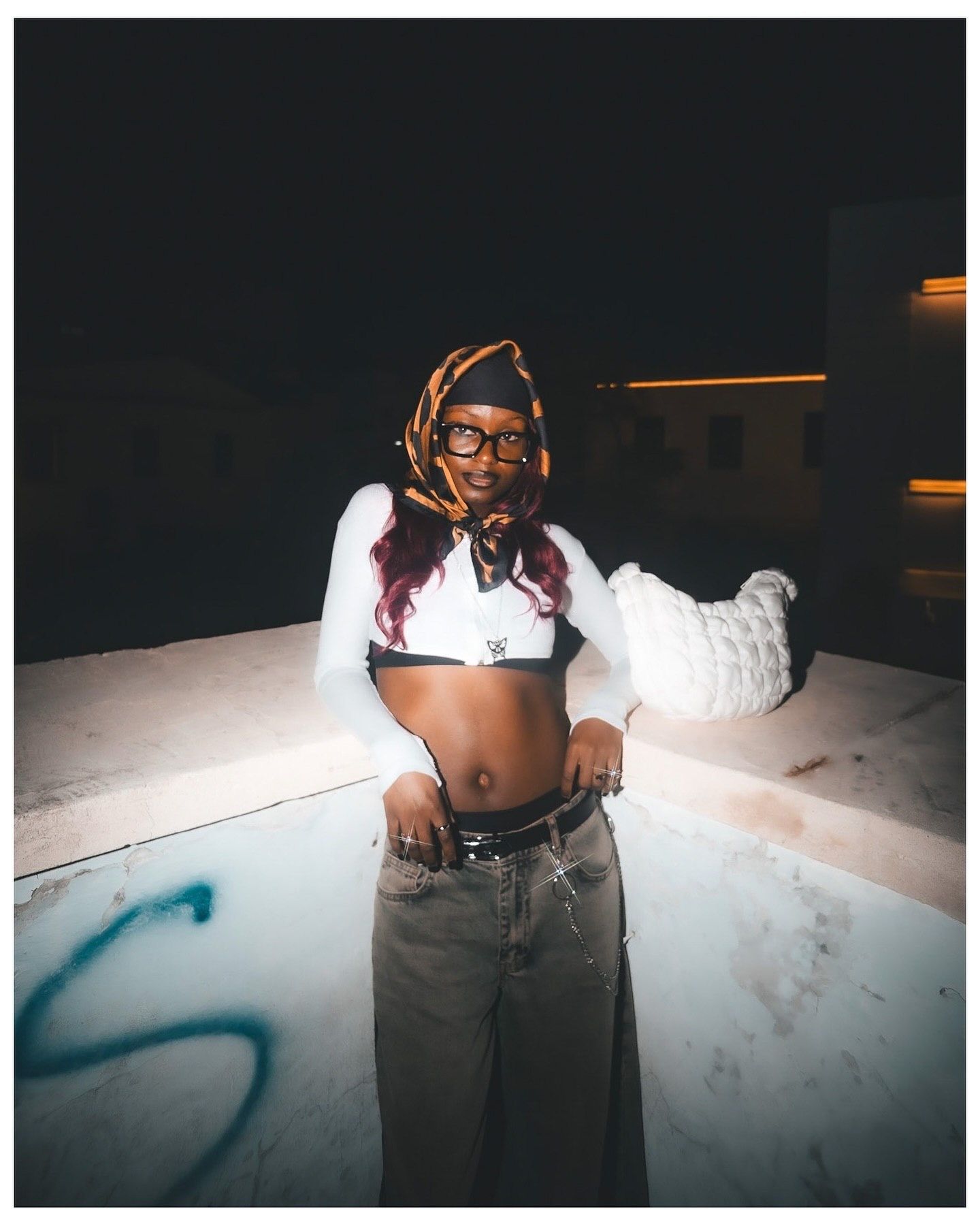 Young woman in crop top, wide-leg jeans, and head scarf poses confidently outdoors at night near graffiti wall.