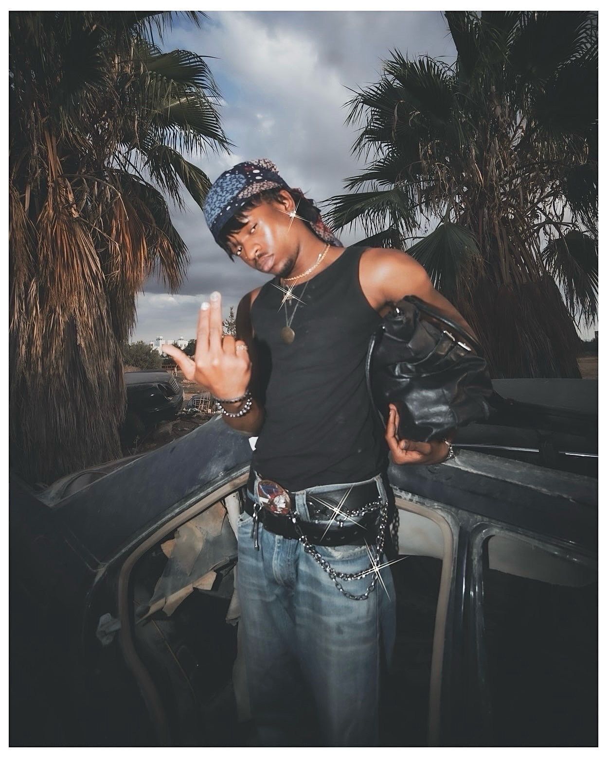 Young man in black tank top, blue bandana, jeans, and chain belt poses near car with palm trees backdrop.