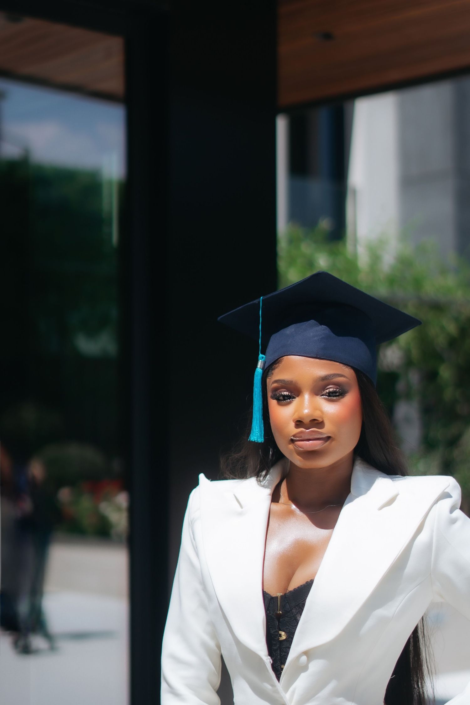 Young Black woman wearing navy graduation cap and white blazer poses in bright sunlight.