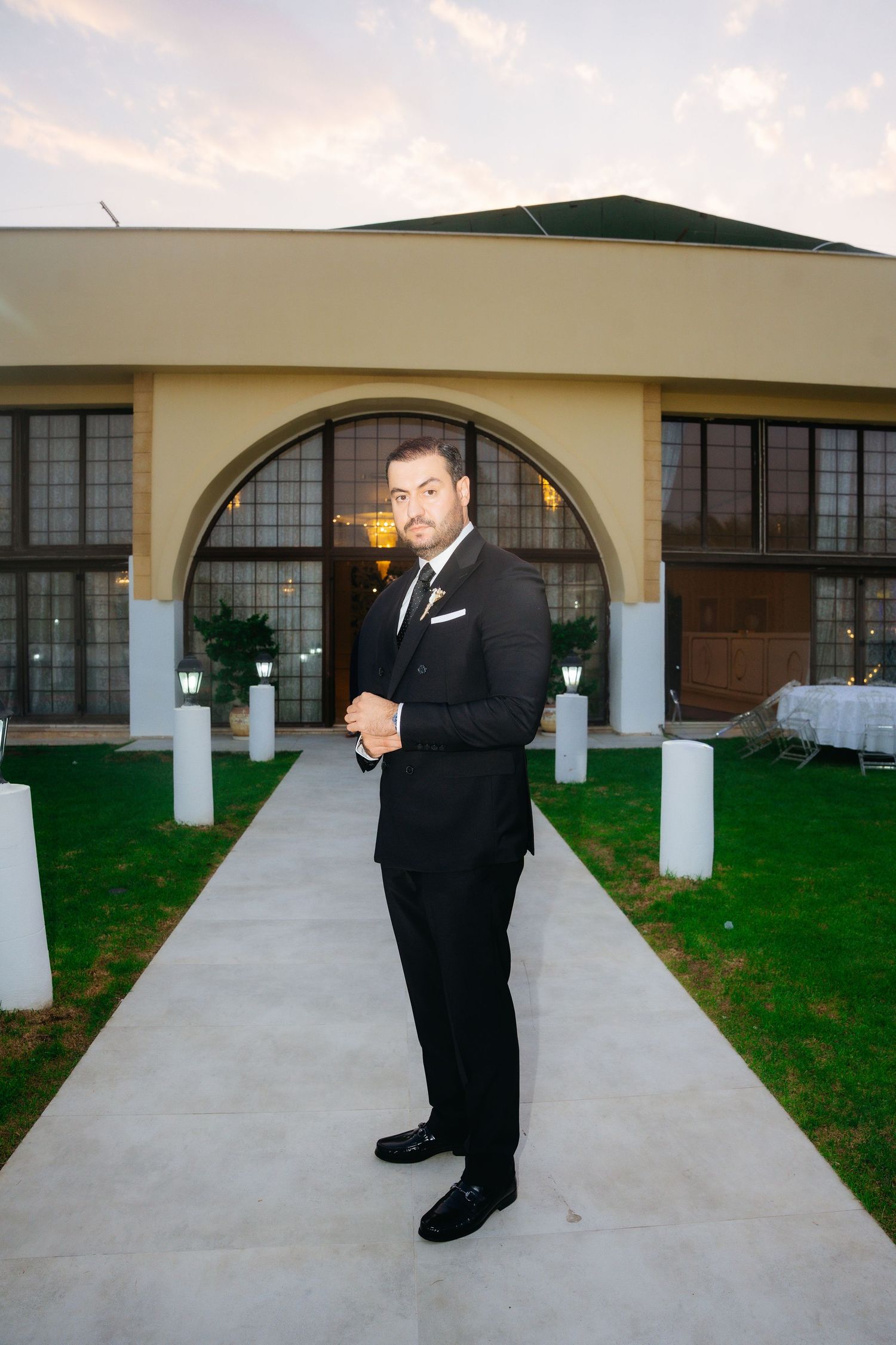 Elegant man in black tuxedo standing on pathway outside luxury venue at dusk.