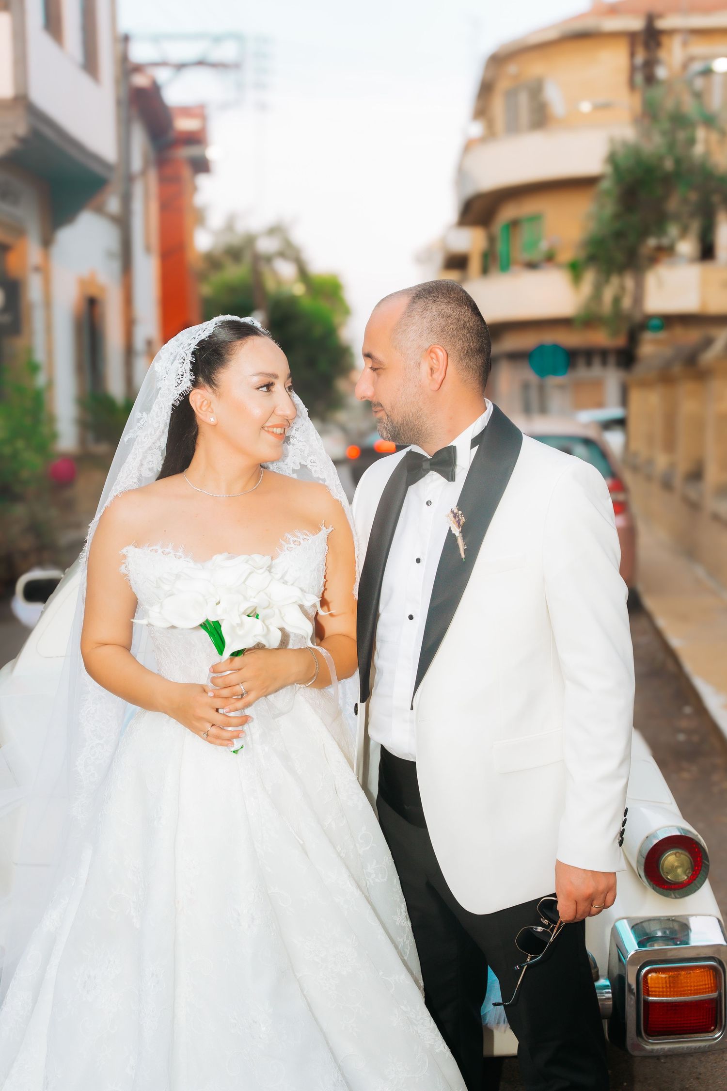 Bride and groom in wedding attire smile at each other on a sunny street, bride holding white bouquet.