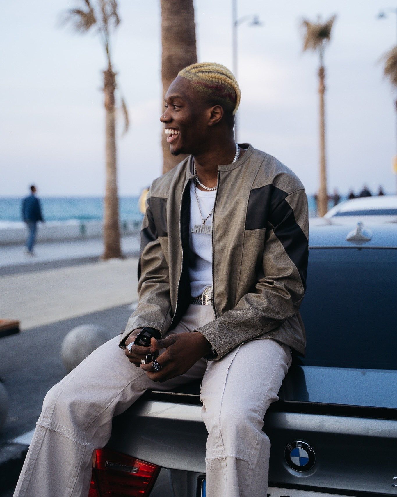 Young man with blonde-tipped hair sits smiling on a BMW car near a beach, wearing a grey jacket and white pants.