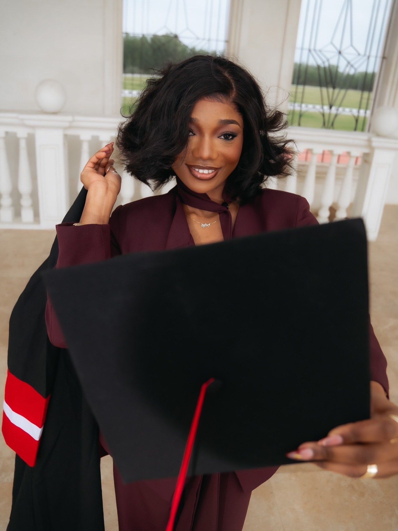 Smiling graduate in burgundy suit holds black cap during graduation photo session indoors.