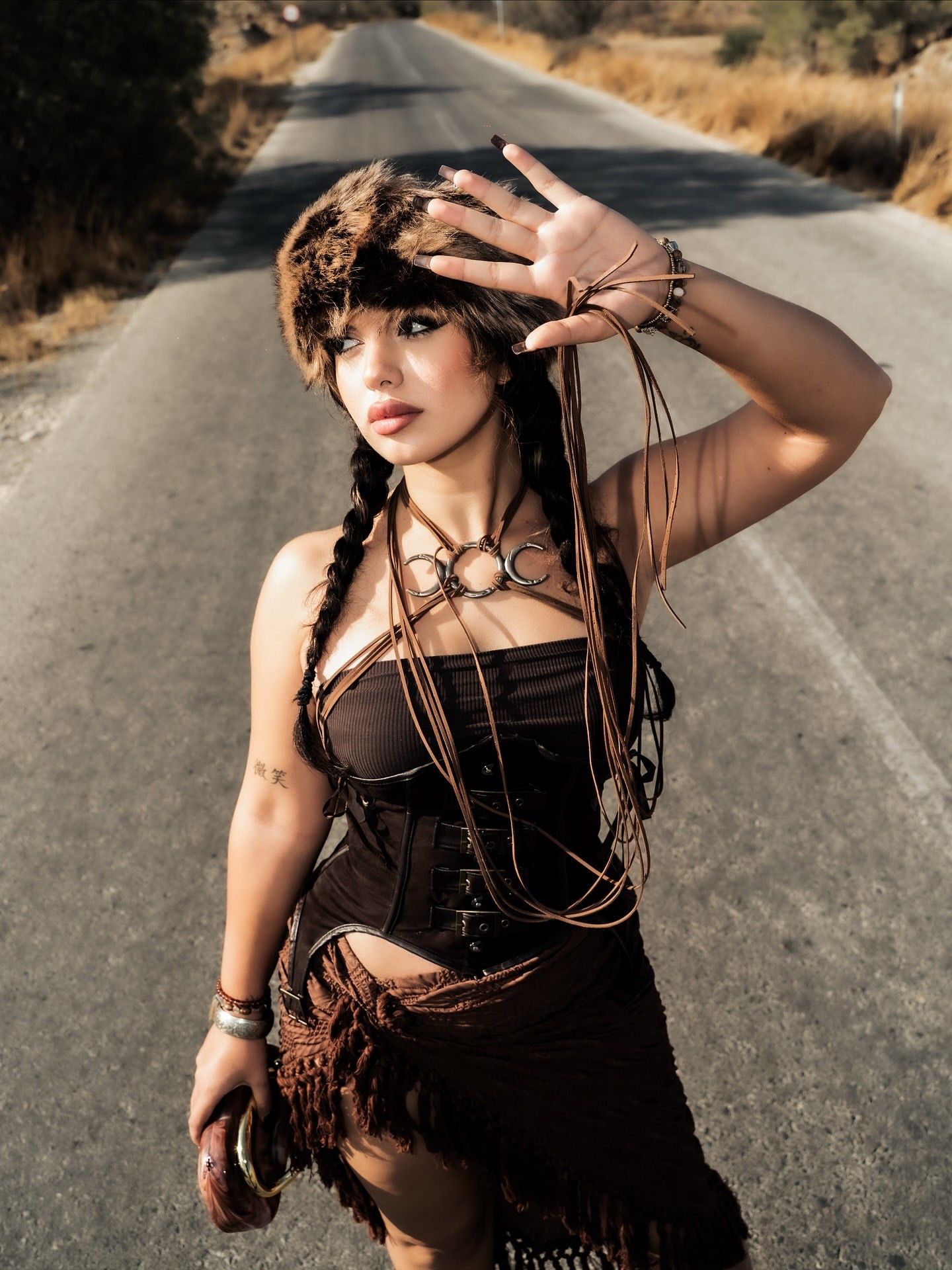 Woman in bohemian costume with fur hat, braids, and brown fringe outfit posing on a rural road.