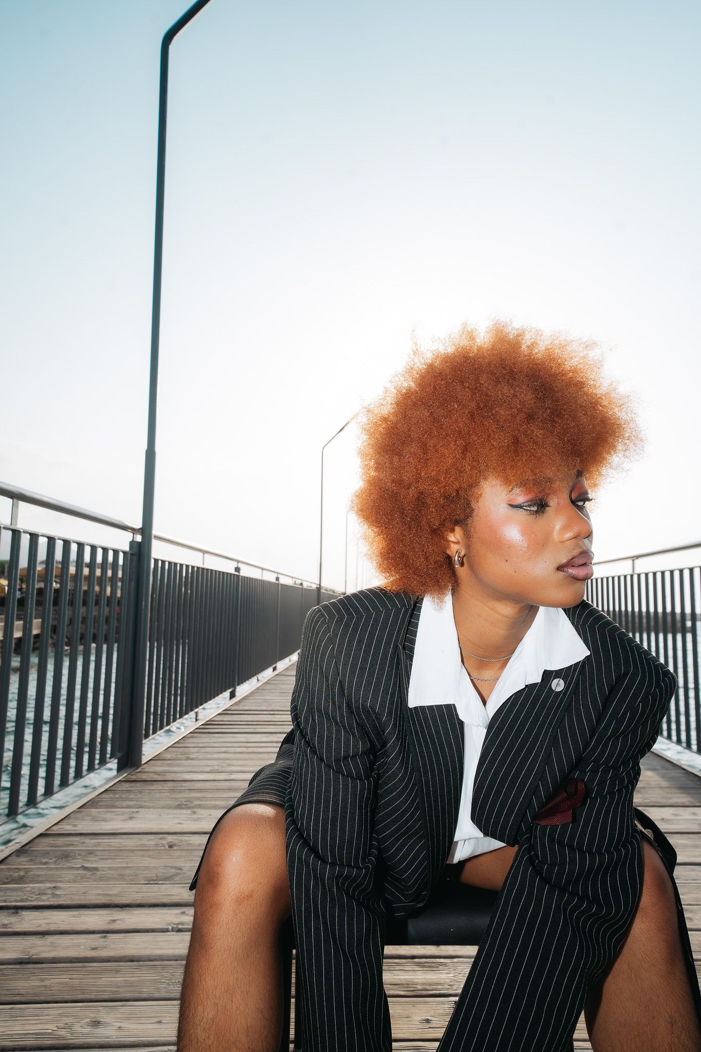 Woman with auburn afro in pinstripe blazer sits pensively on wooden boardwalk by railing.