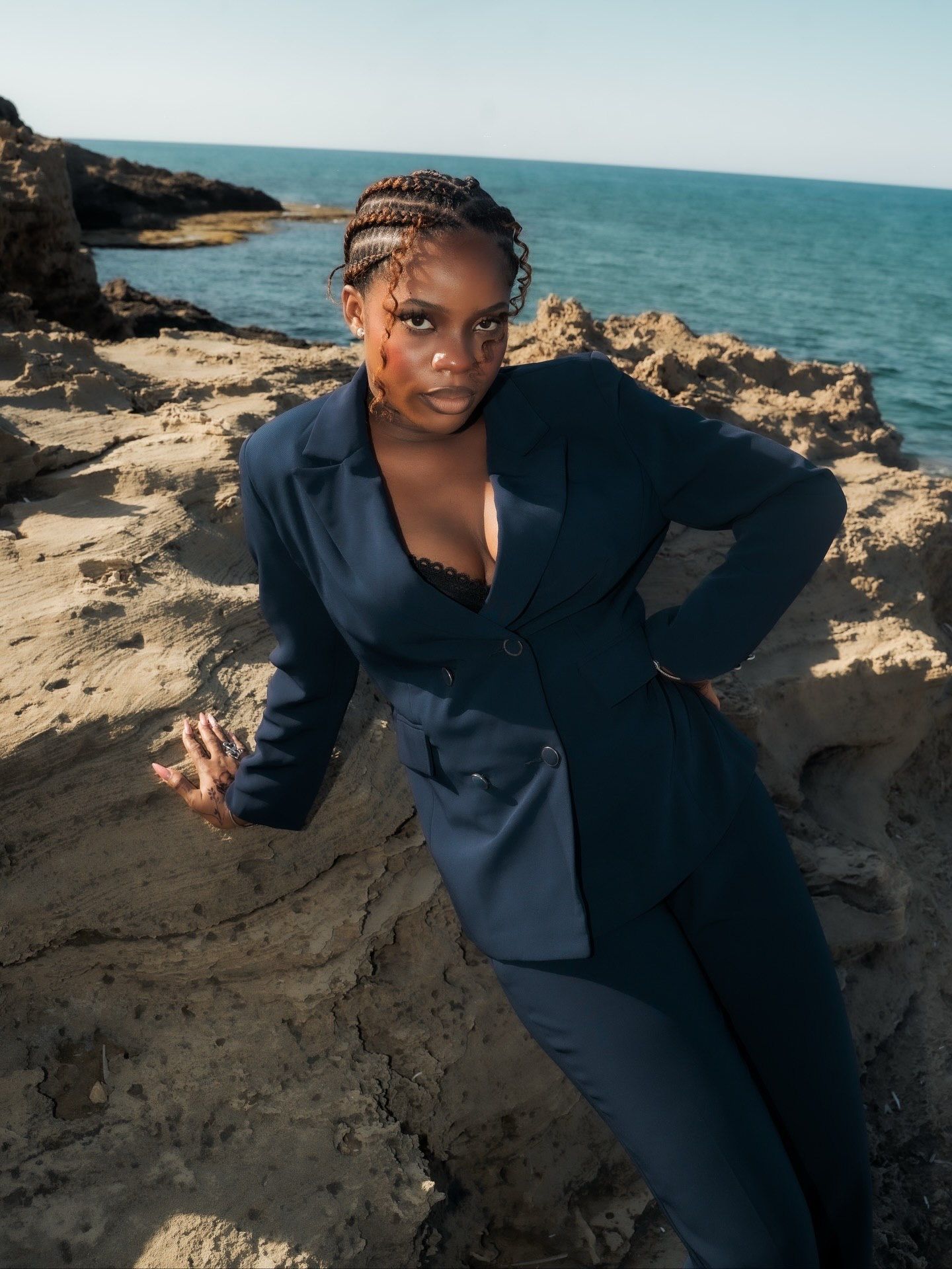 Woman in navy pantsuit poses dramatically on rocky coastal cliffs with turquoise ocean behind.