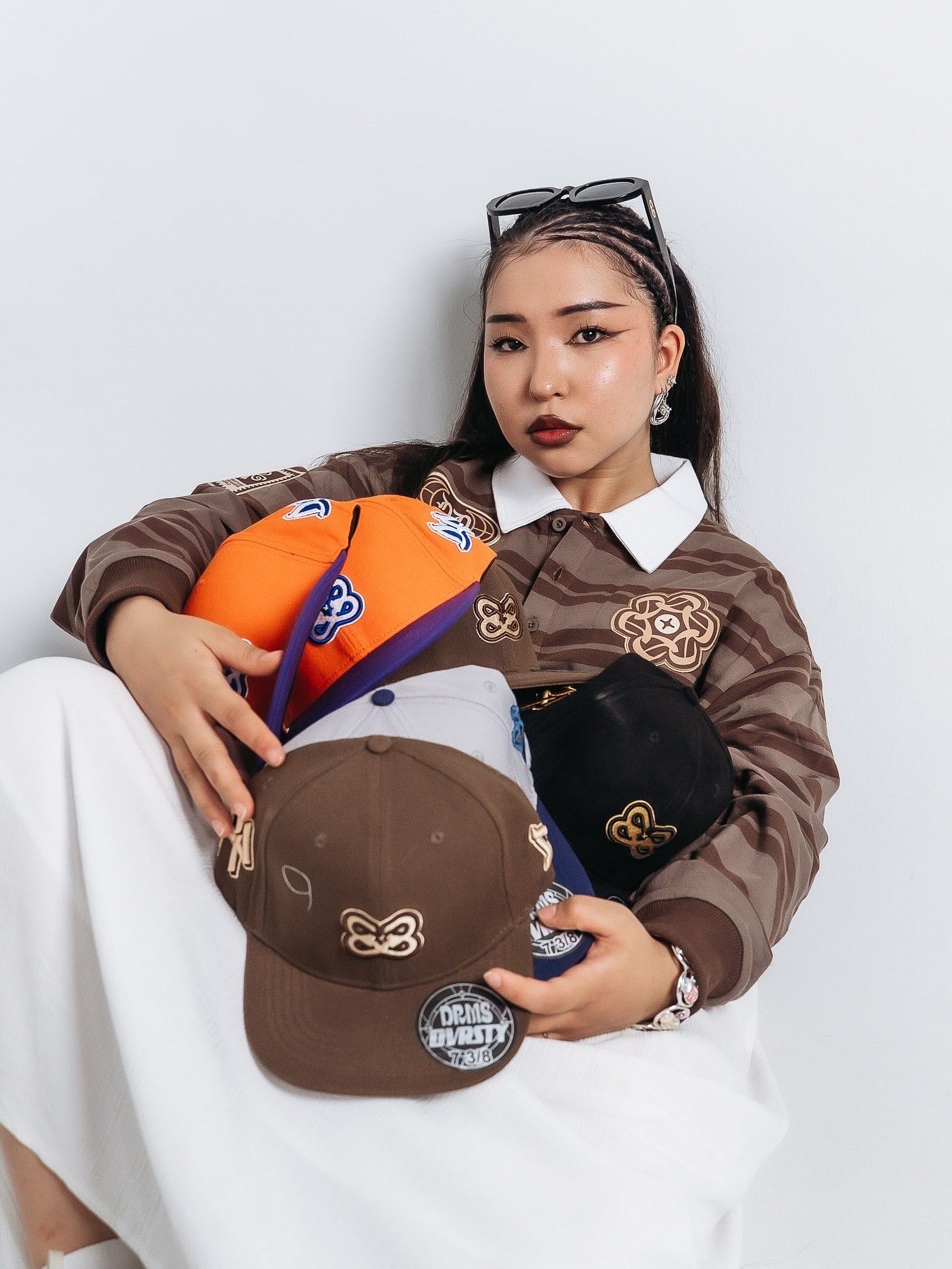 Young woman poses with multiple branded snapback caps in brown, orange, and black, wearing a brown embroidered jacket.
