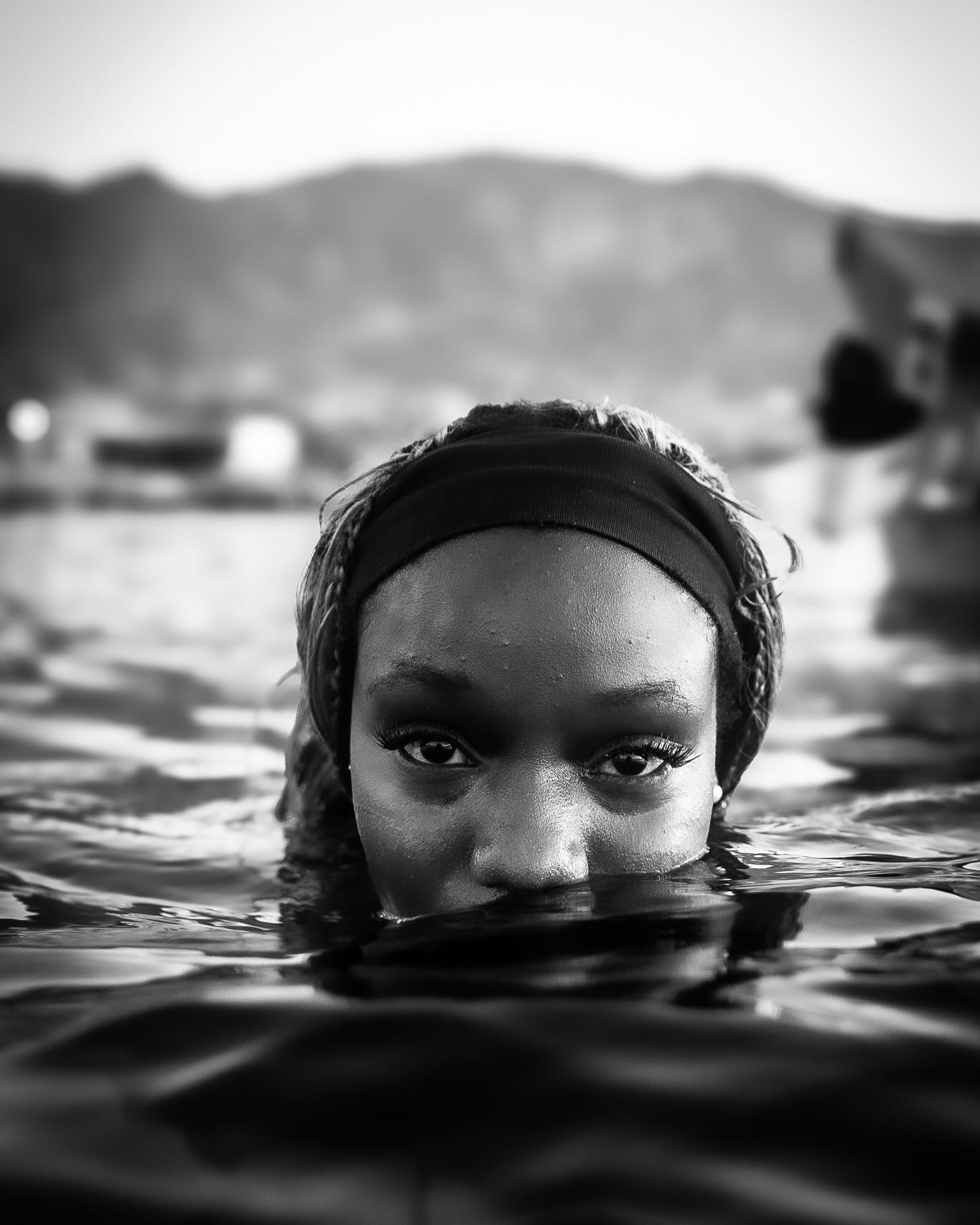 Black and white portrait of a young woman submerged in water up to her nose, with mountains in the background.