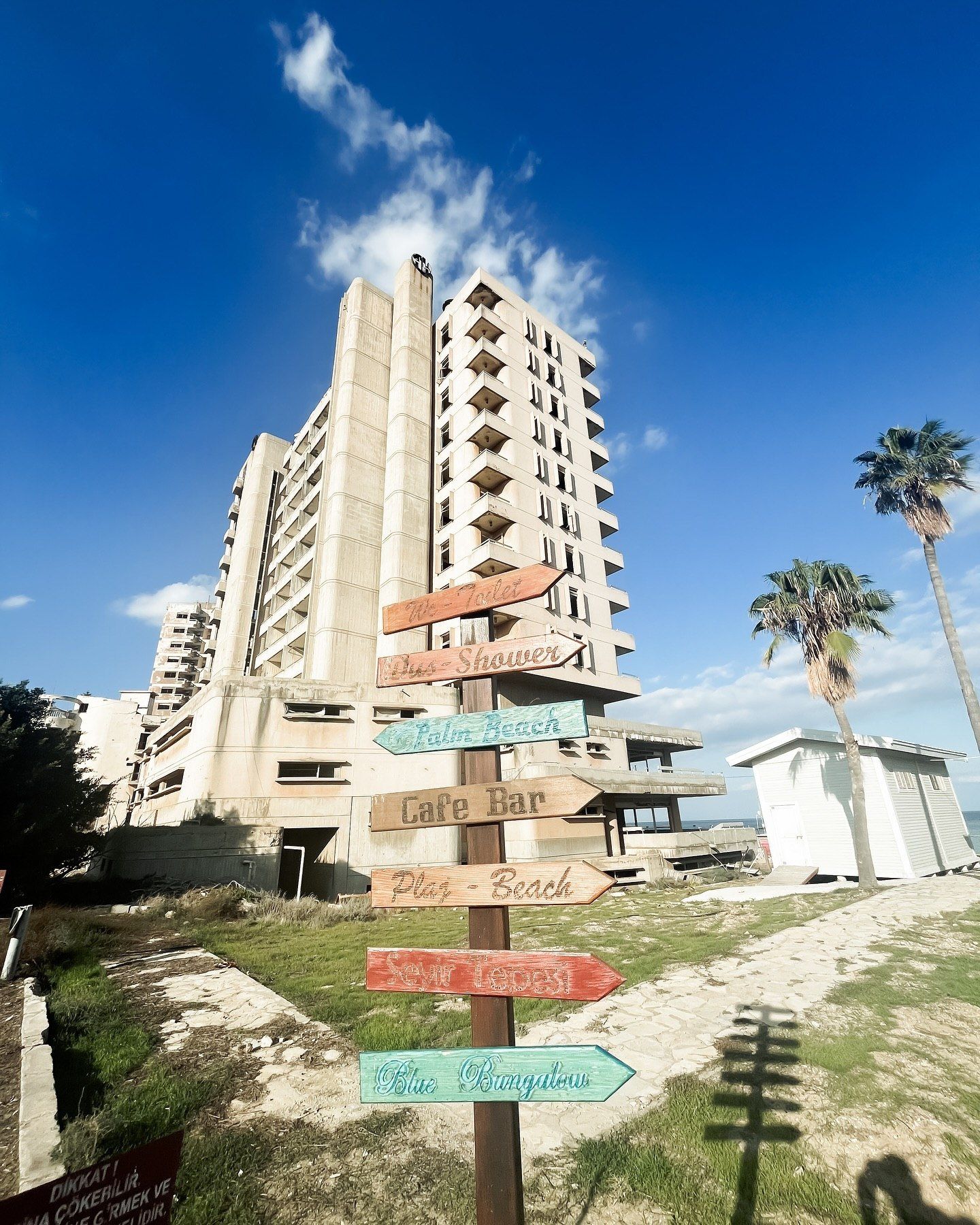 Abandoned hotel tower with colorful directional signs pointing to beach, cafe bar, and showers under a bright blue sky.