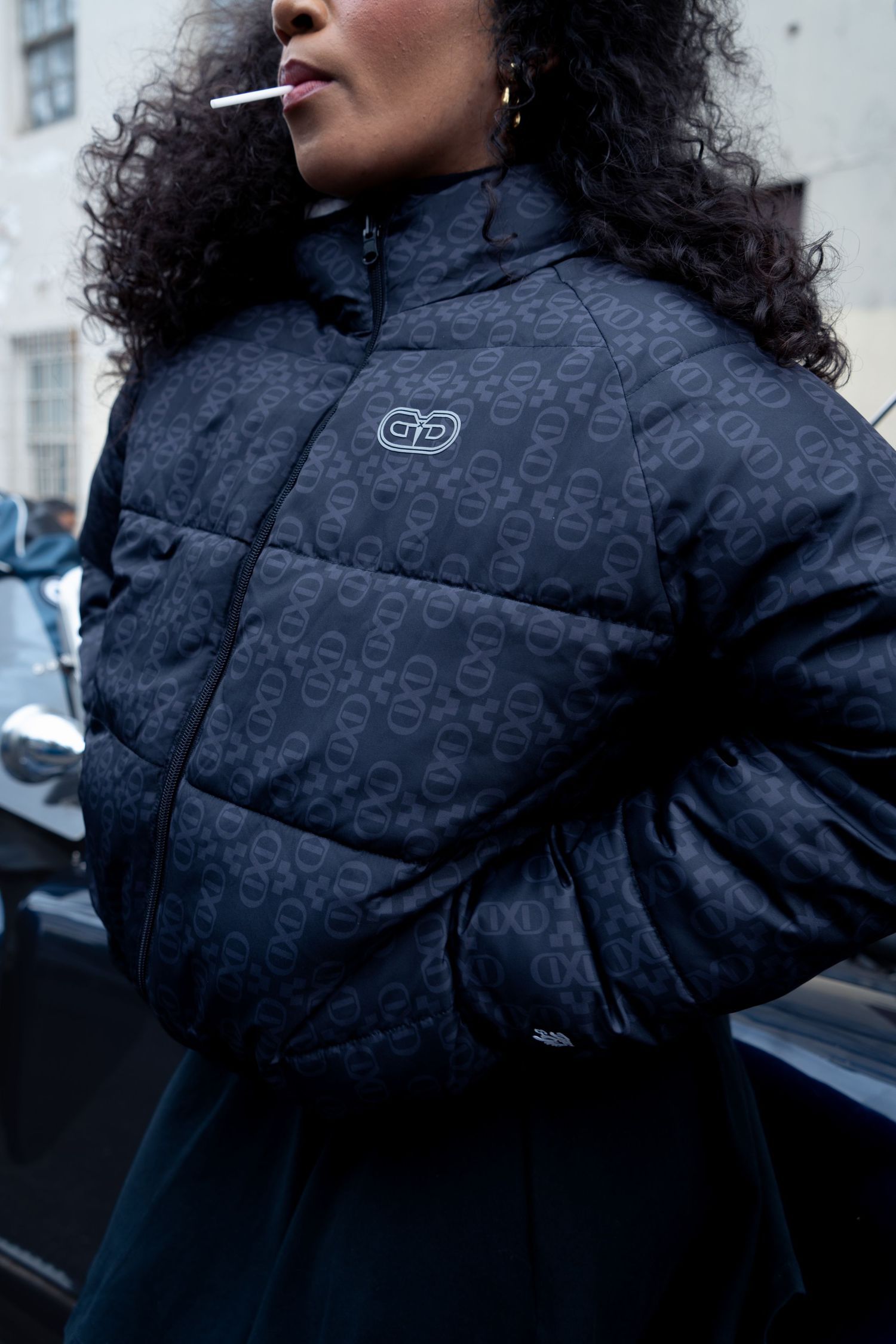 Woman wearing a black quilted puffer jacket with monogram pattern and logo, smoking a cigarette outdoors.