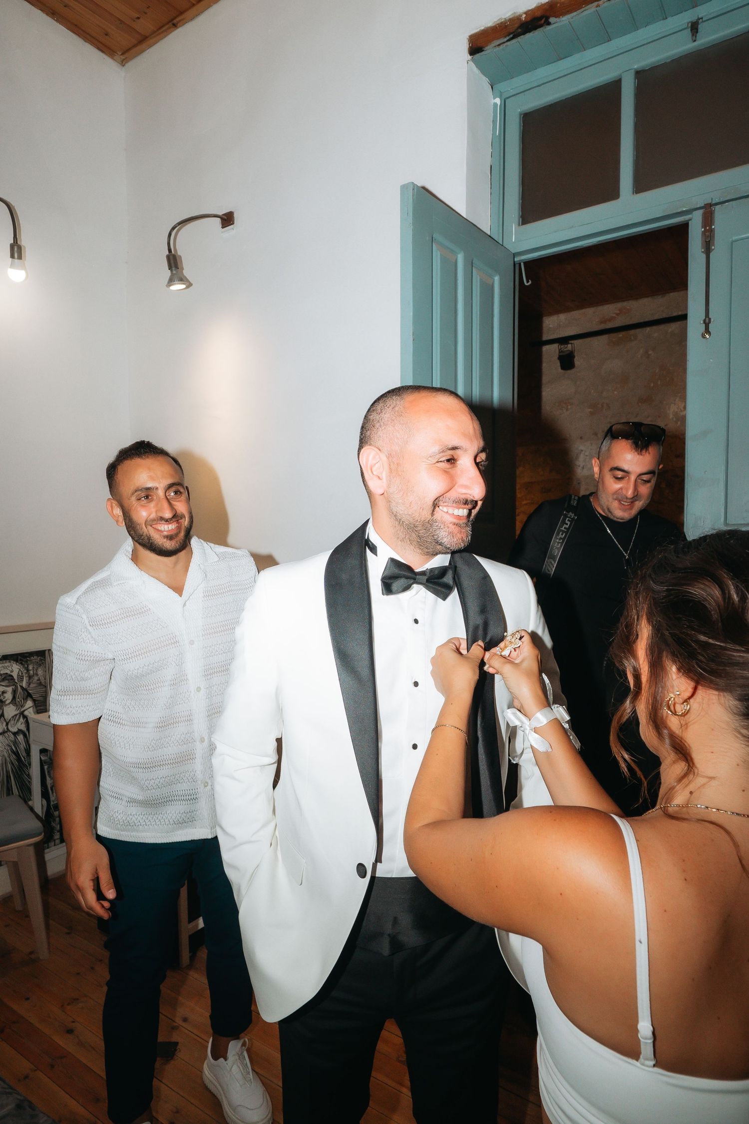 Groom in white tuxedo smiles as bride adjusts his boutonniere at wedding reception with guests in background.