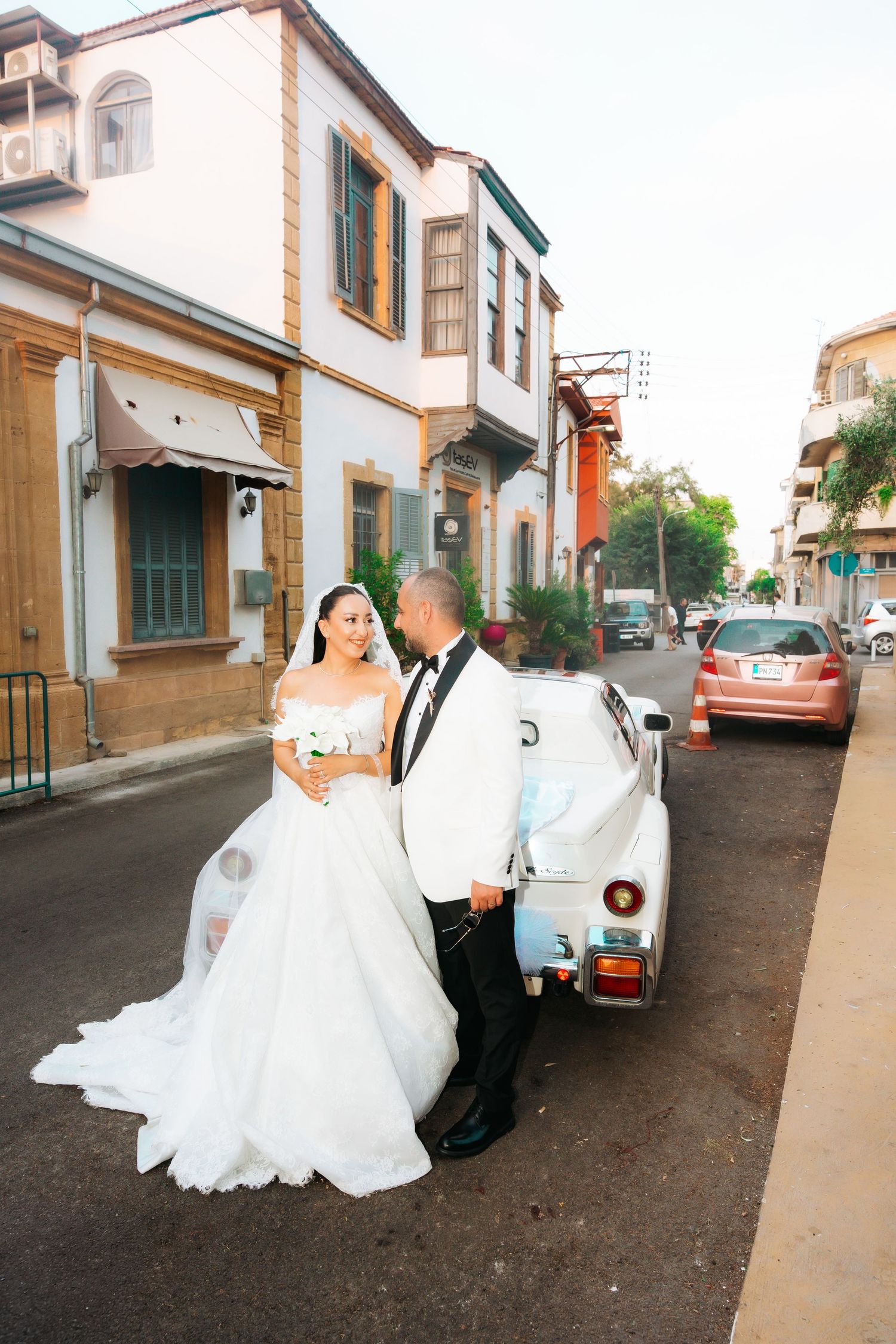 Bride in white gown and groom in white tuxedo pose beside a vintage car on a charming street.