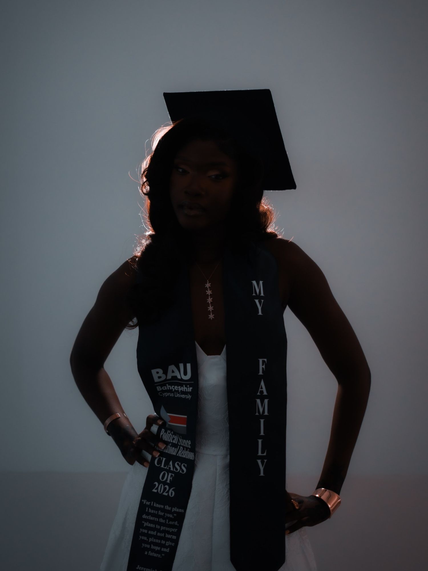 Graduate in cap and gown with 'My Family' stole stands in dramatic shadow lighting.