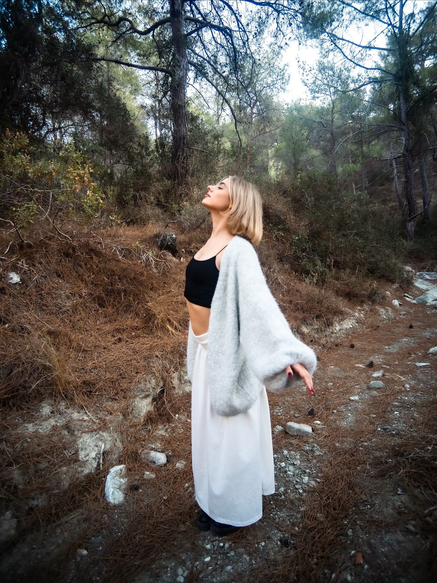 Blonde woman in white maxi cardigan and black crop top posing on a forest trail with trees in background.