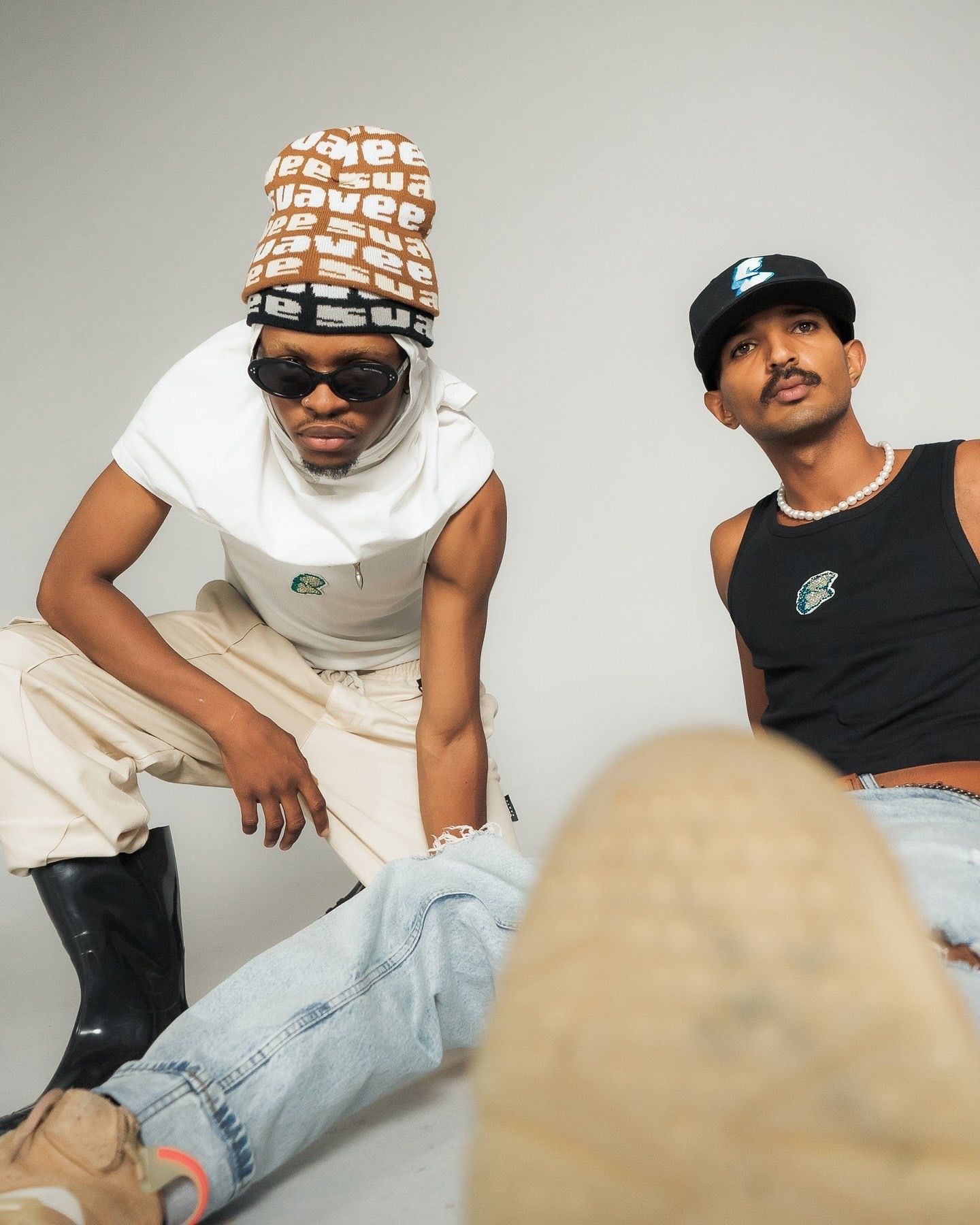 Two stylish young men posing in streetwear, one wearing a beanie and sunglasses, the other in a black cap and tank top.