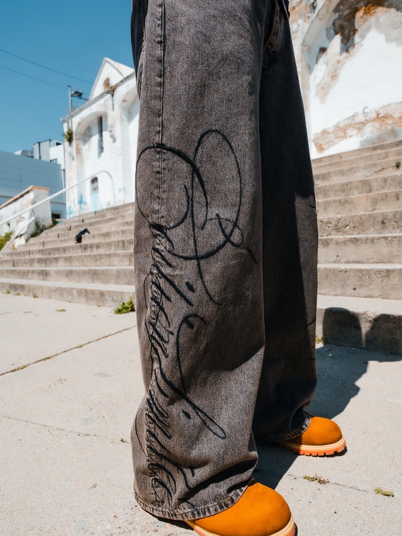 Wide-leg dark wash jeans with black script graffiti print, styled with orange slides on a sunny urban sidewalk.