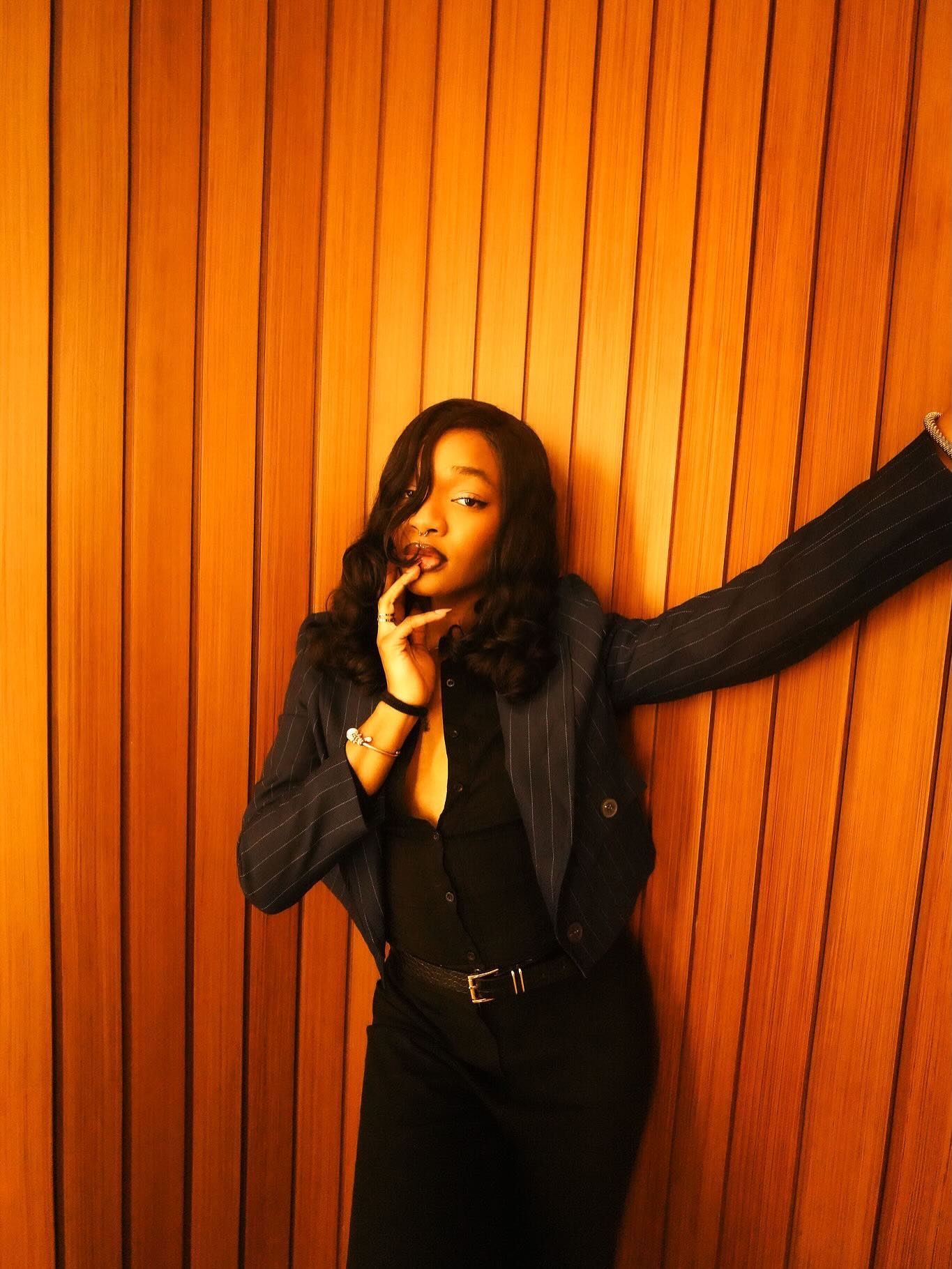 A stylish young woman in a navy pinstripe blazer and black outfit poses confidently against a warm wooden panel wall.