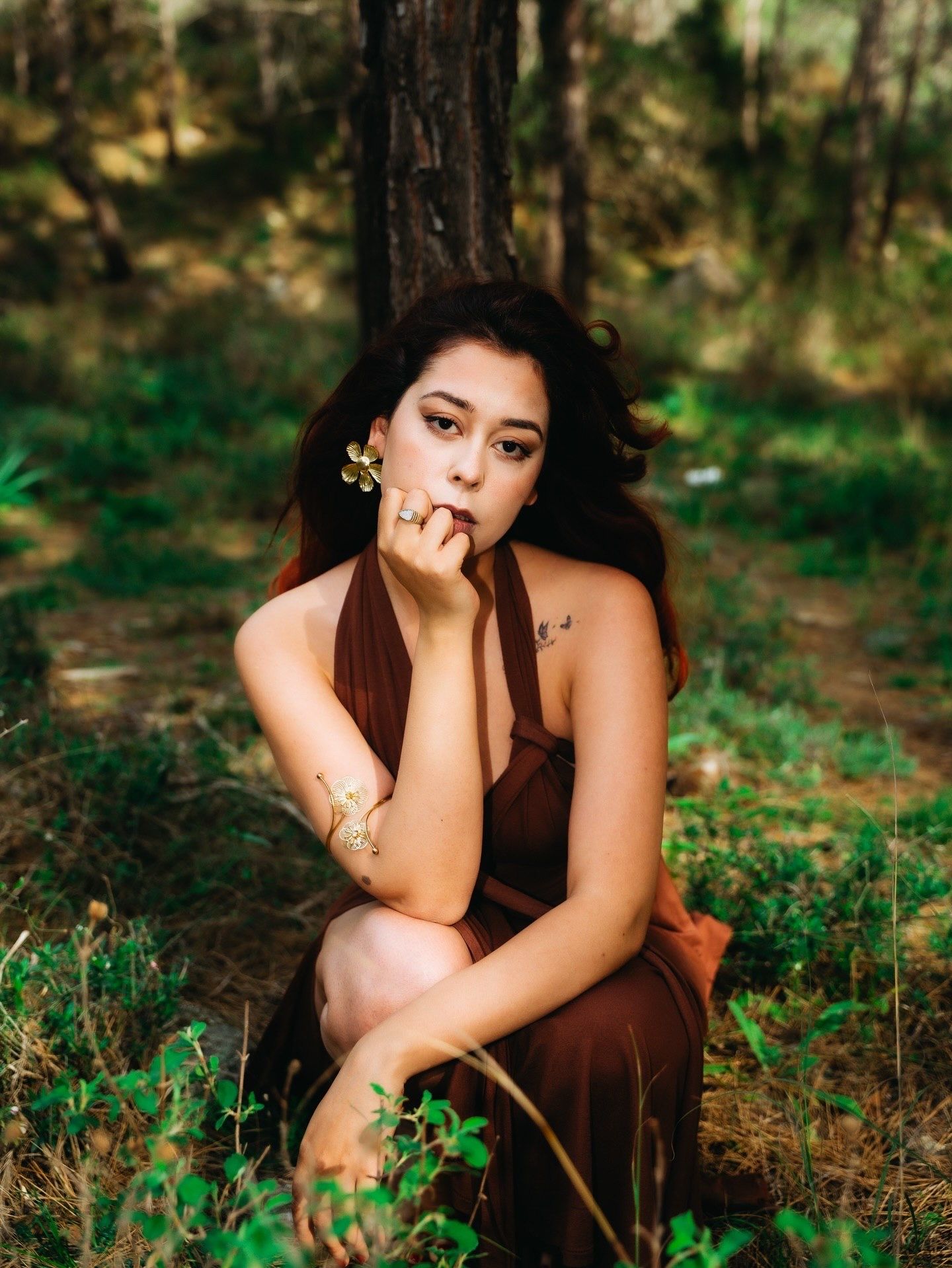 Young woman in brown dress sitting by tree in forest, holding a small flower near her face.