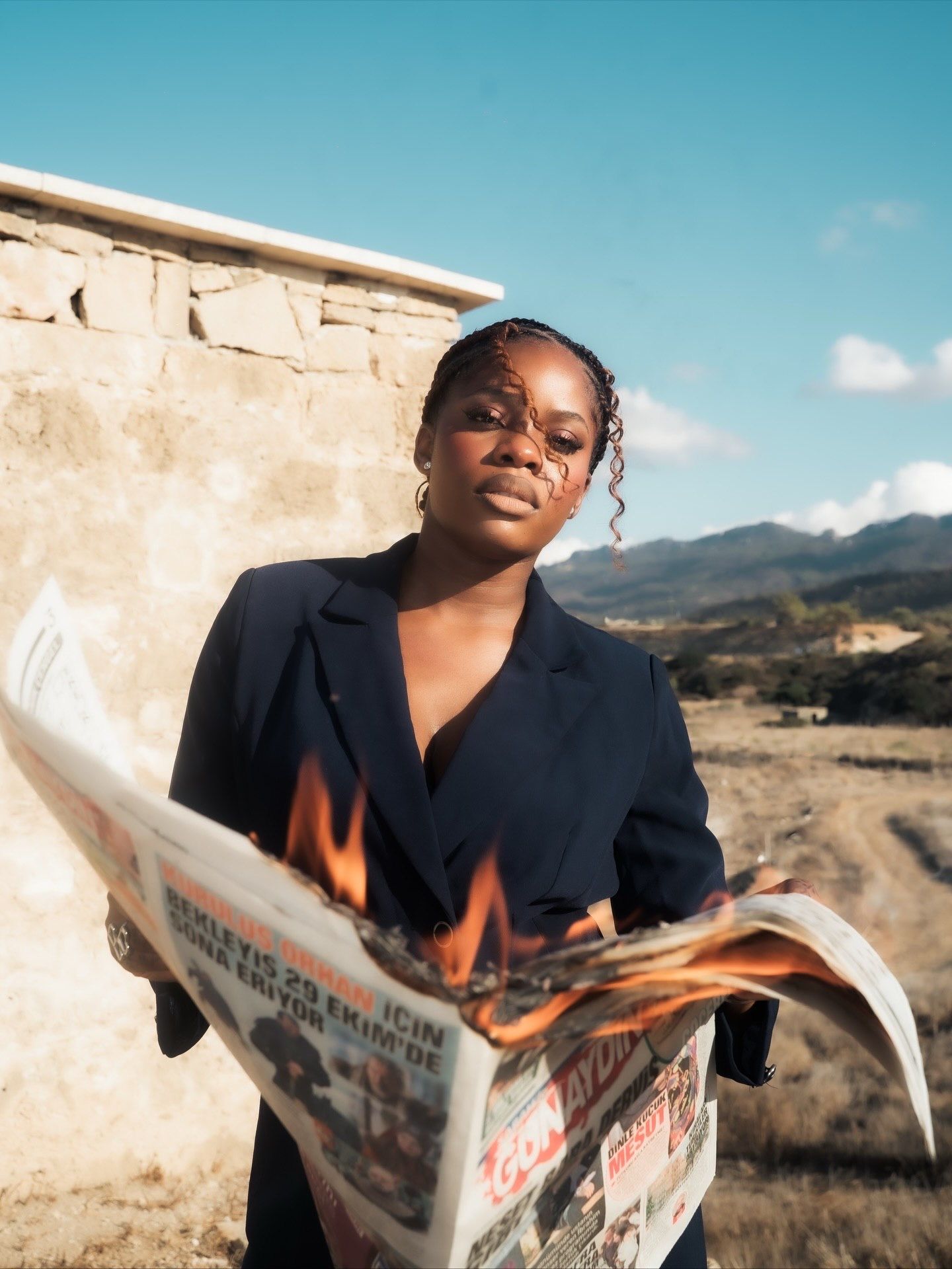 Black woman in navy blazer holds burning newspaper outdoors with mountains and blue sky in background.