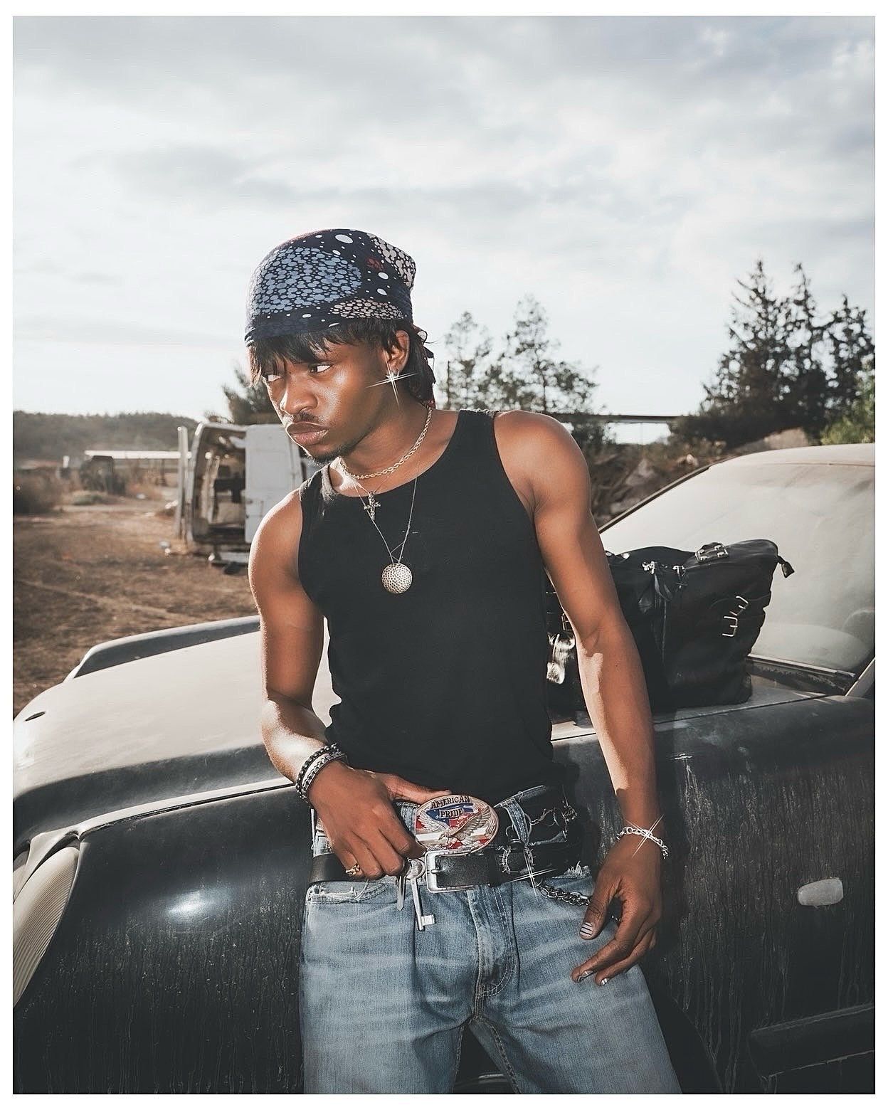 Young man in black tank top, bandana, jeans, and chain necklace leaning on a dark car outdoors.