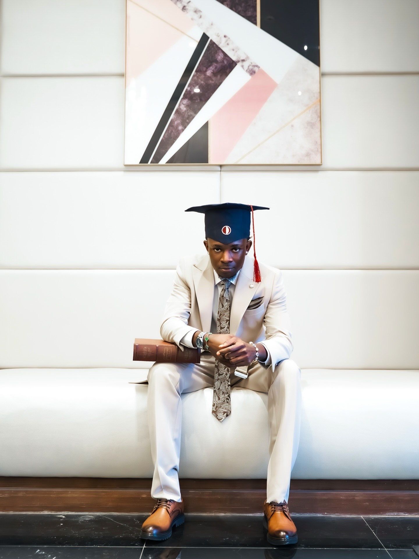 Young man in cream suit and graduation cap sits pensively on white bench indoors.