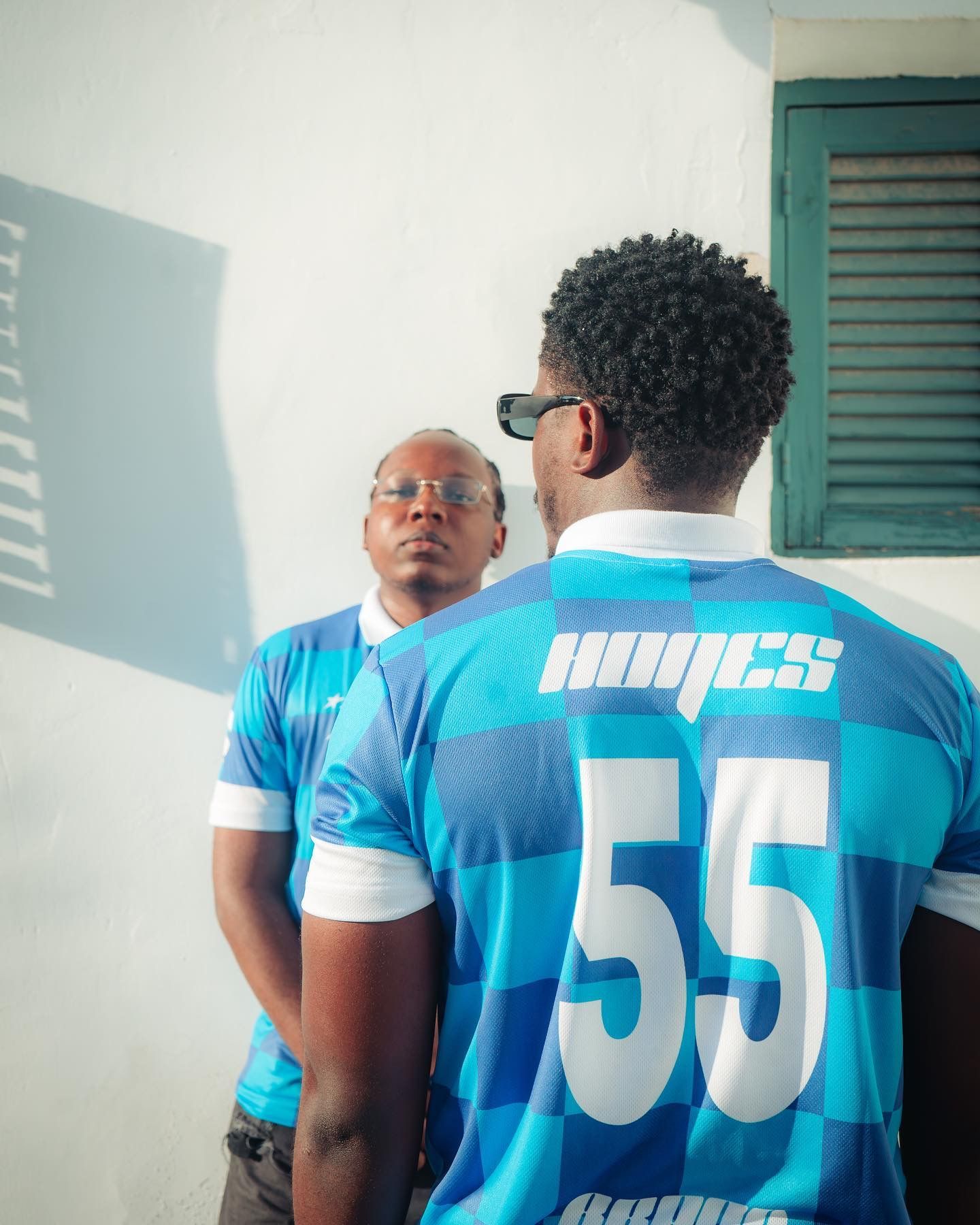 Two men wearing blue striped jerseys numbered 55 stand outdoors near a white wall and green window.