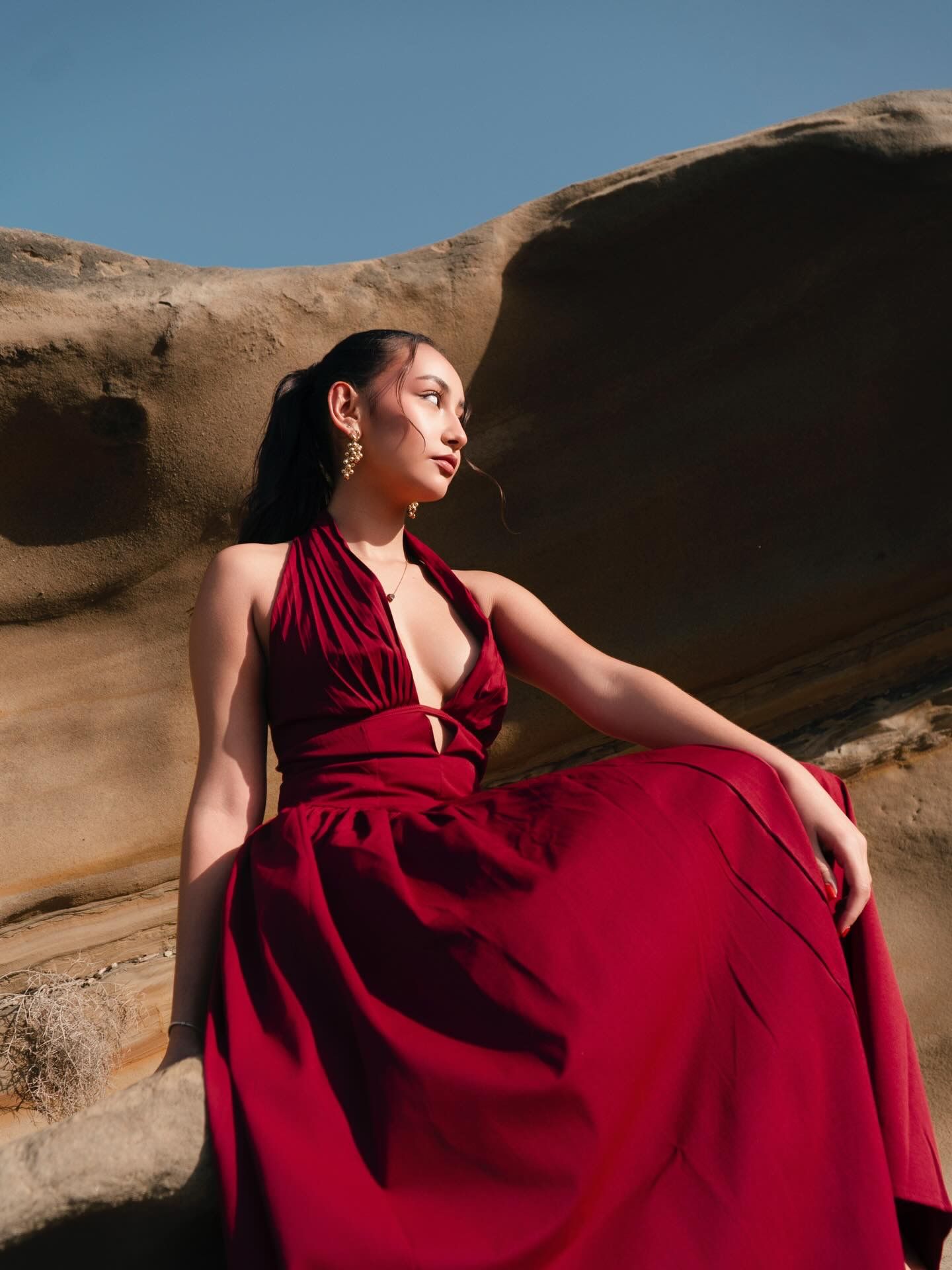 Woman in red halter maxi dress posed on sandstone rocks under blue sky.