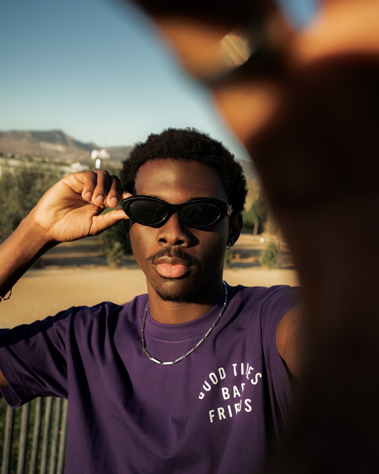 Young man in purple Good Times Bad Friends tee and black sunglasses taking a selfie outdoors on a sunny day.
