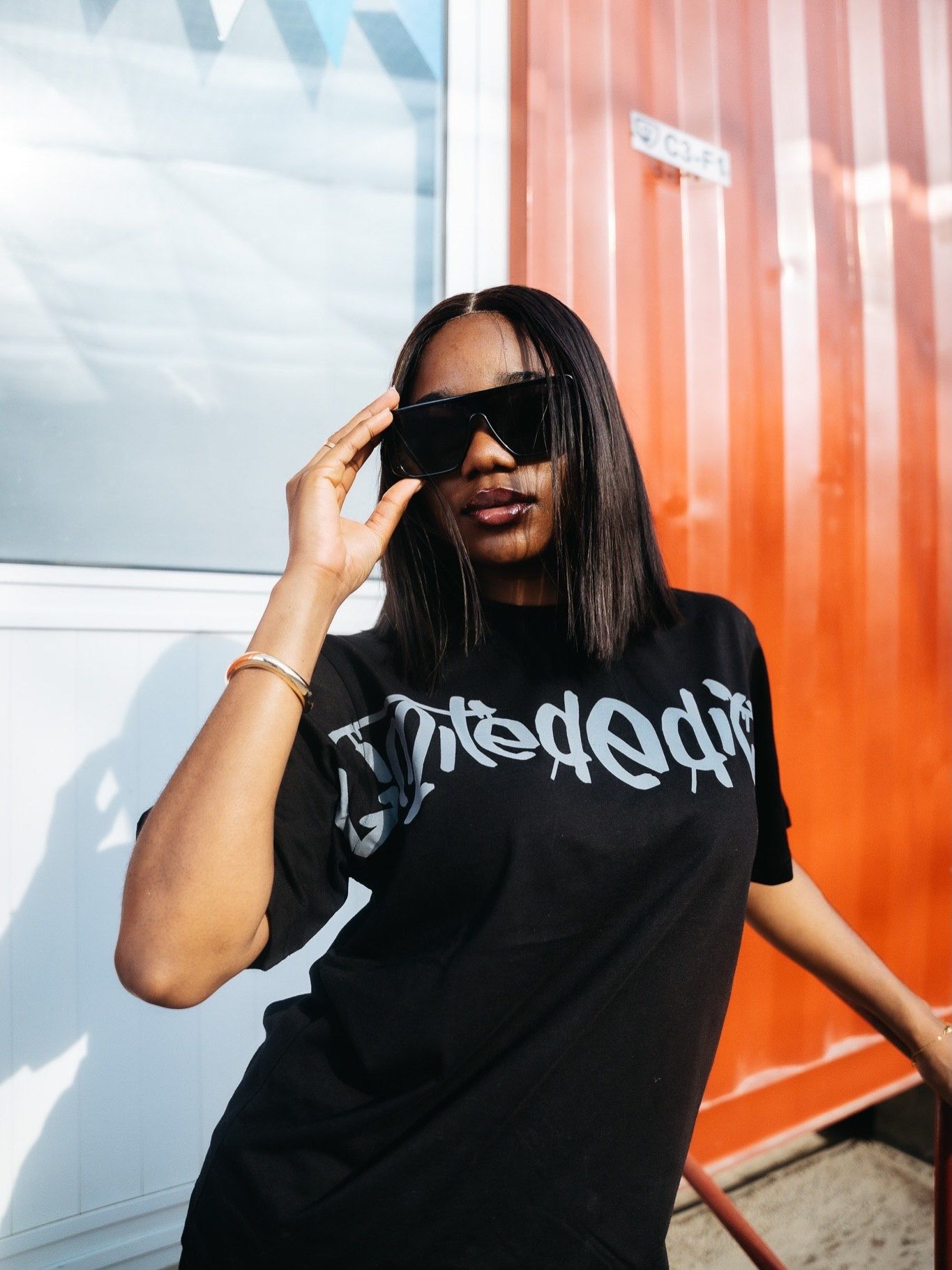 Woman in black graphic tee and oversized sunglasses posing outdoors near a red wall in sunlight.