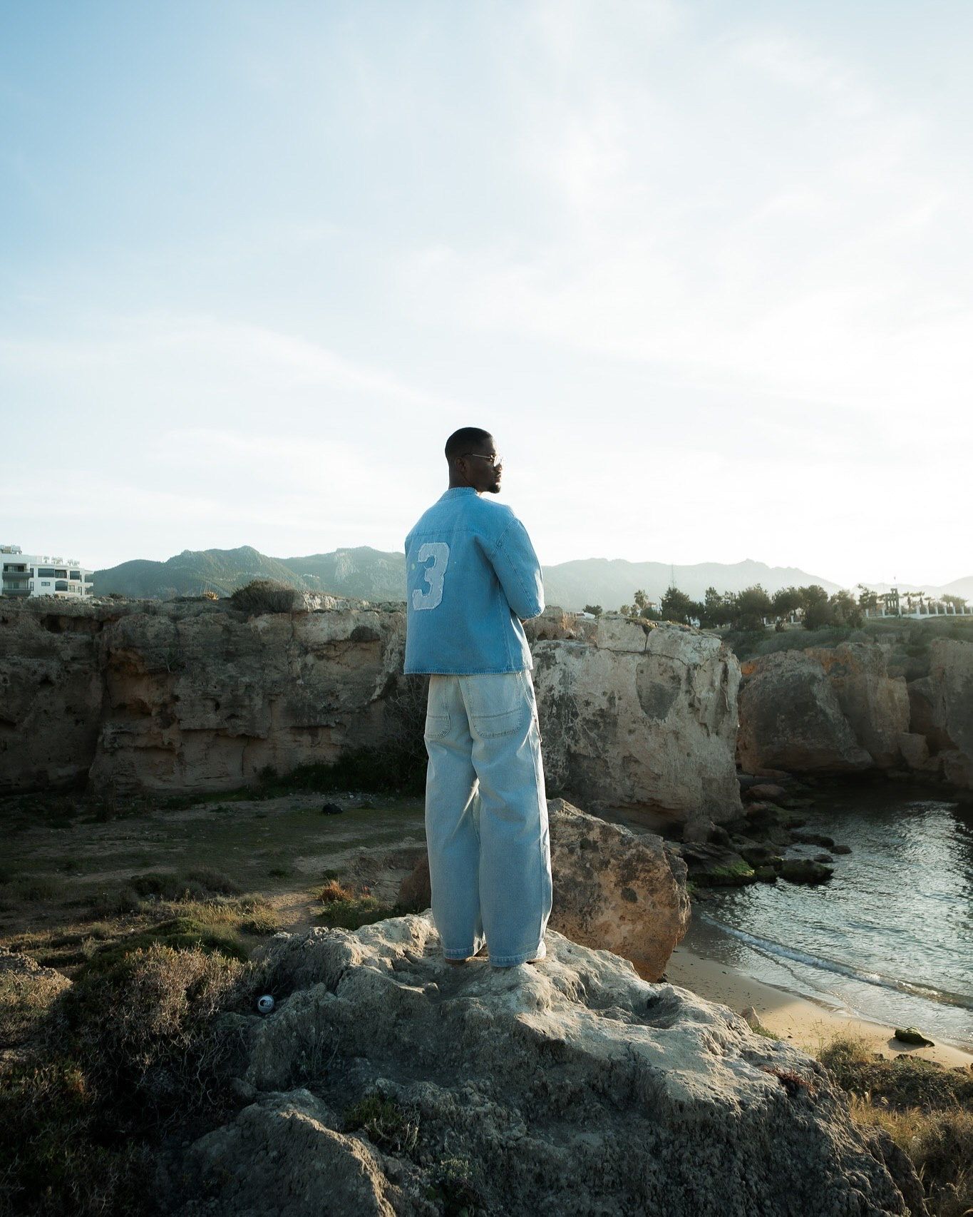 A man in a blue sweater and light pants stands on rocky coastal cliffs overlooking a serene cove with mountains beyond.