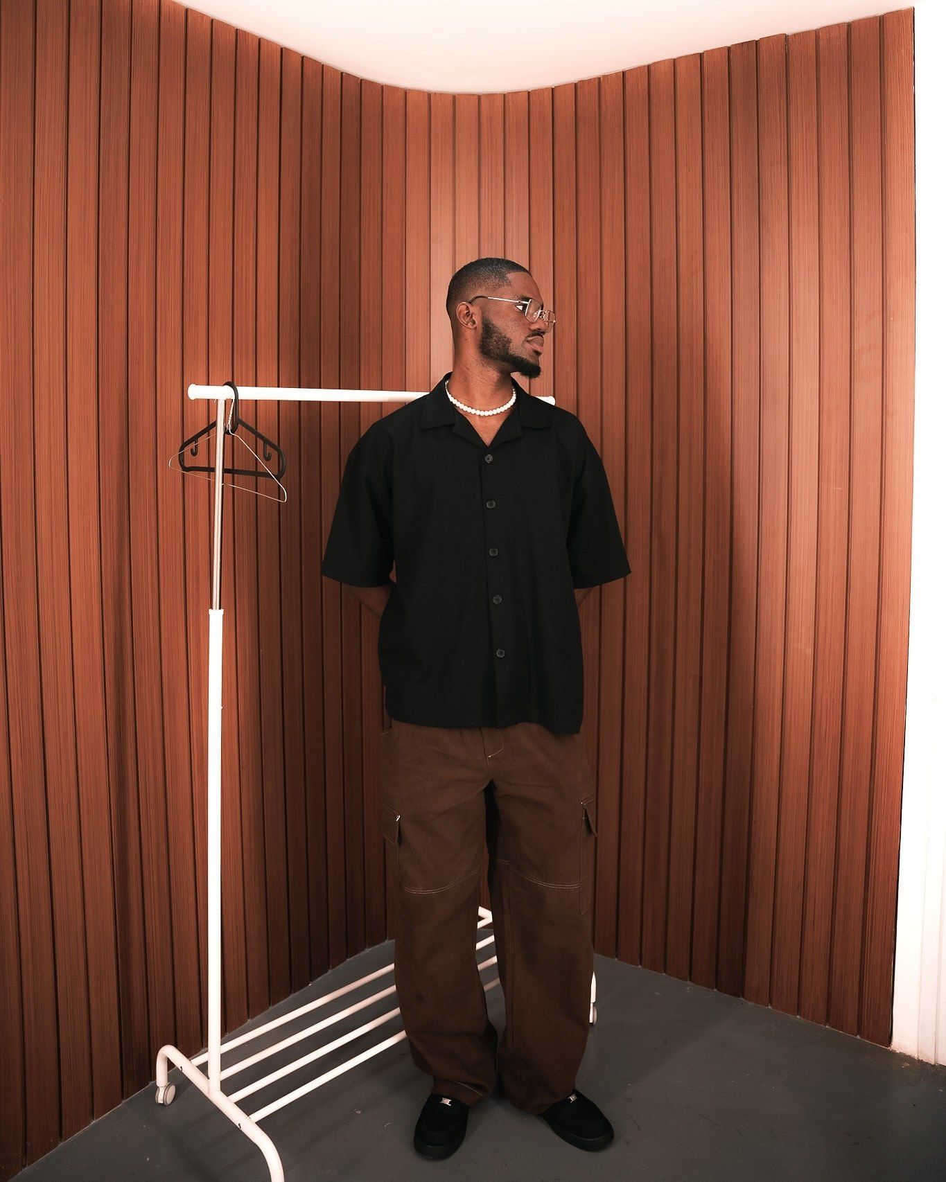 Man in black shirt and brown cargo pants stands by a clothing rack against a striped terracotta wall.