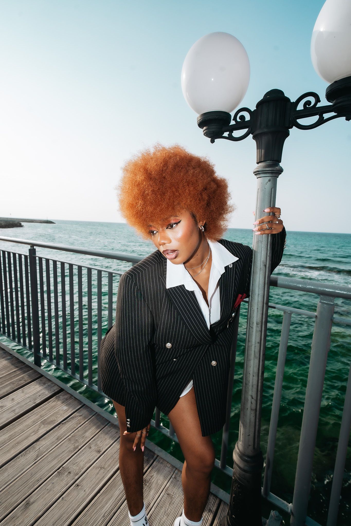 Stylish woman with red afro leans on pier lamp post wearing oversized pinstripe blazer by ocean.
