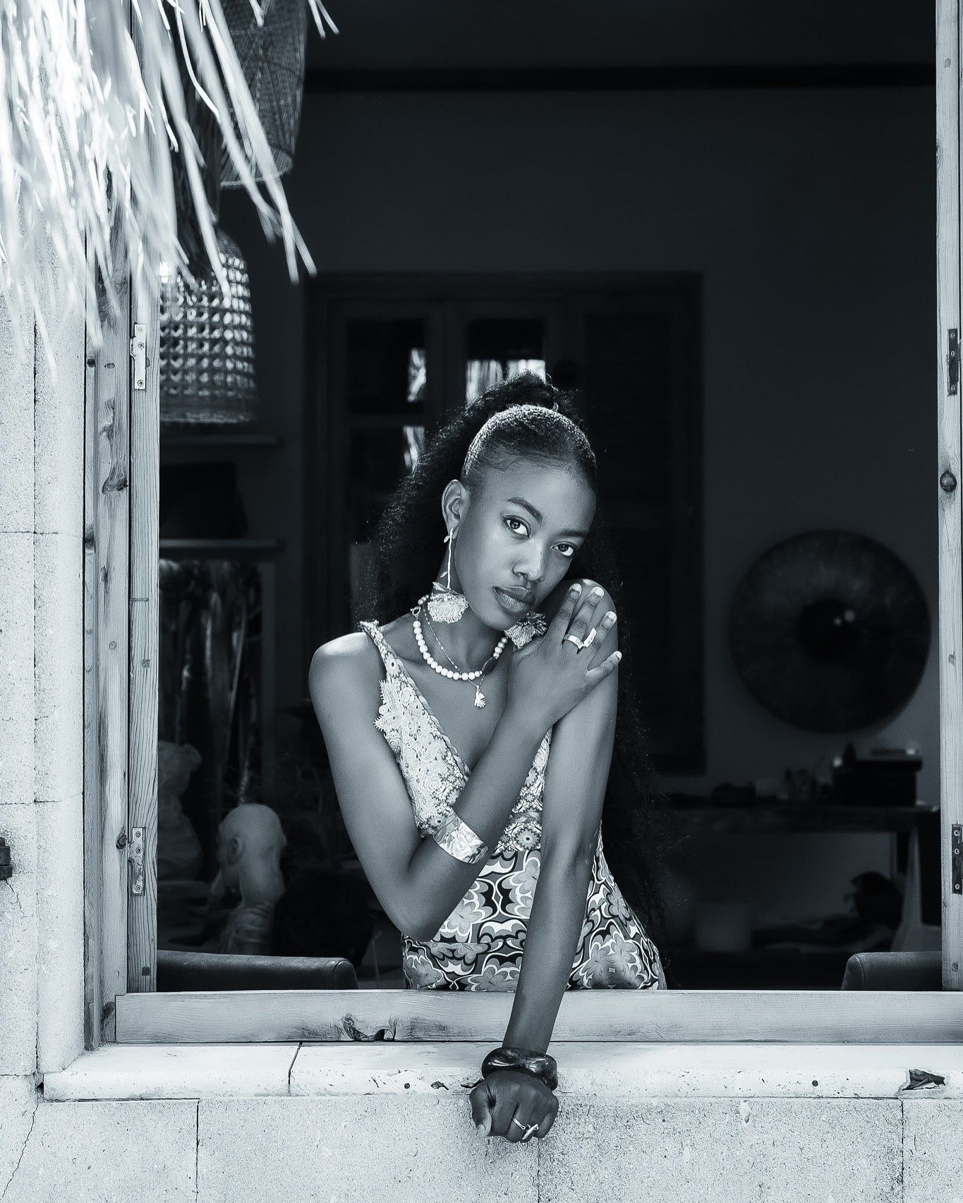 Black and white portrait of a young woman leaning on a windowsill, wearing jewelry and a floral dress.