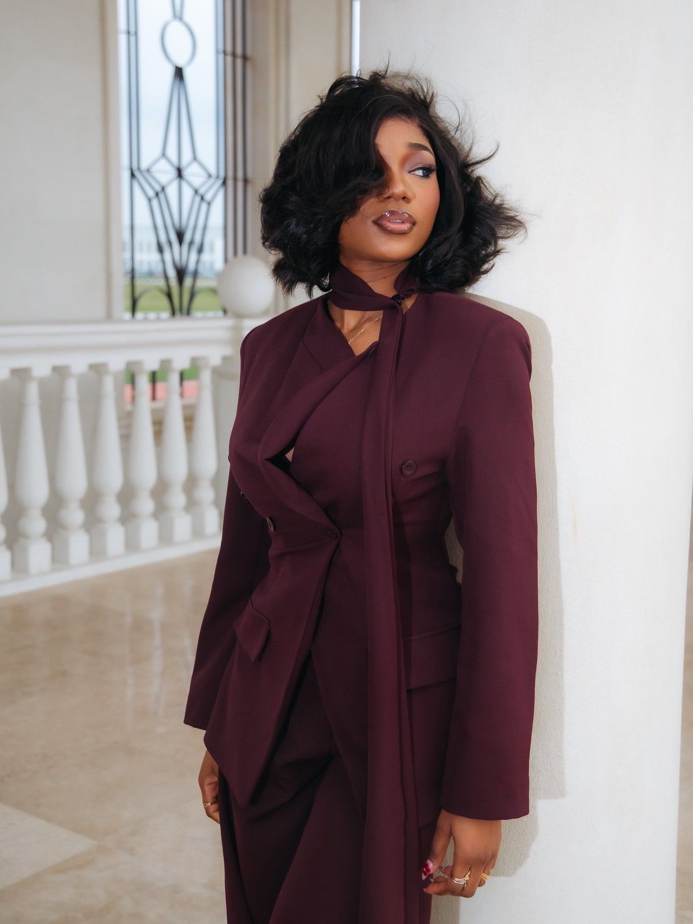Stylish woman in burgundy wrap suit poses confidently near white balustrade in bright interior.