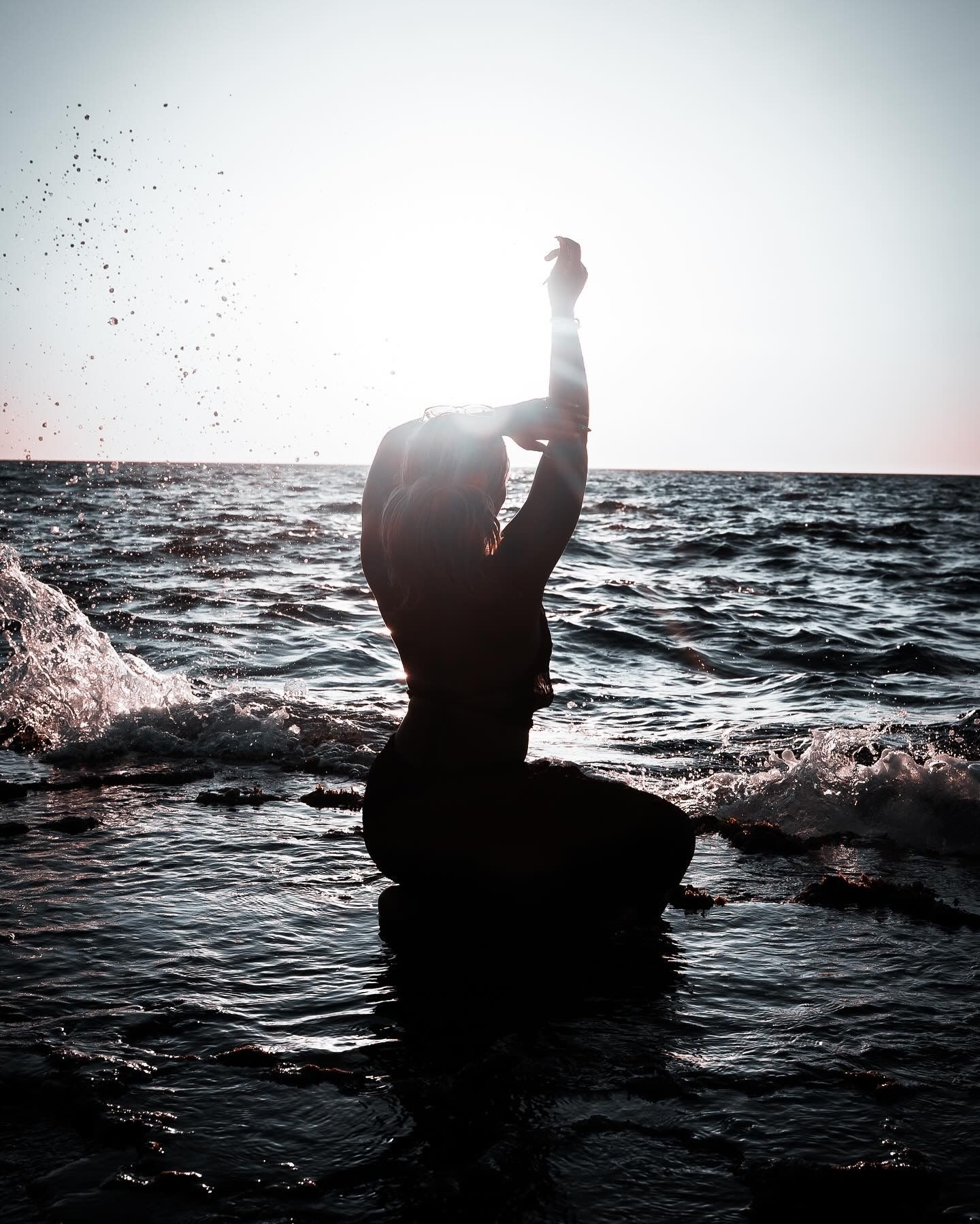 Silhouette of a person kneeling in ocean waves with arms raised, backlit by a glowing sunset sky.