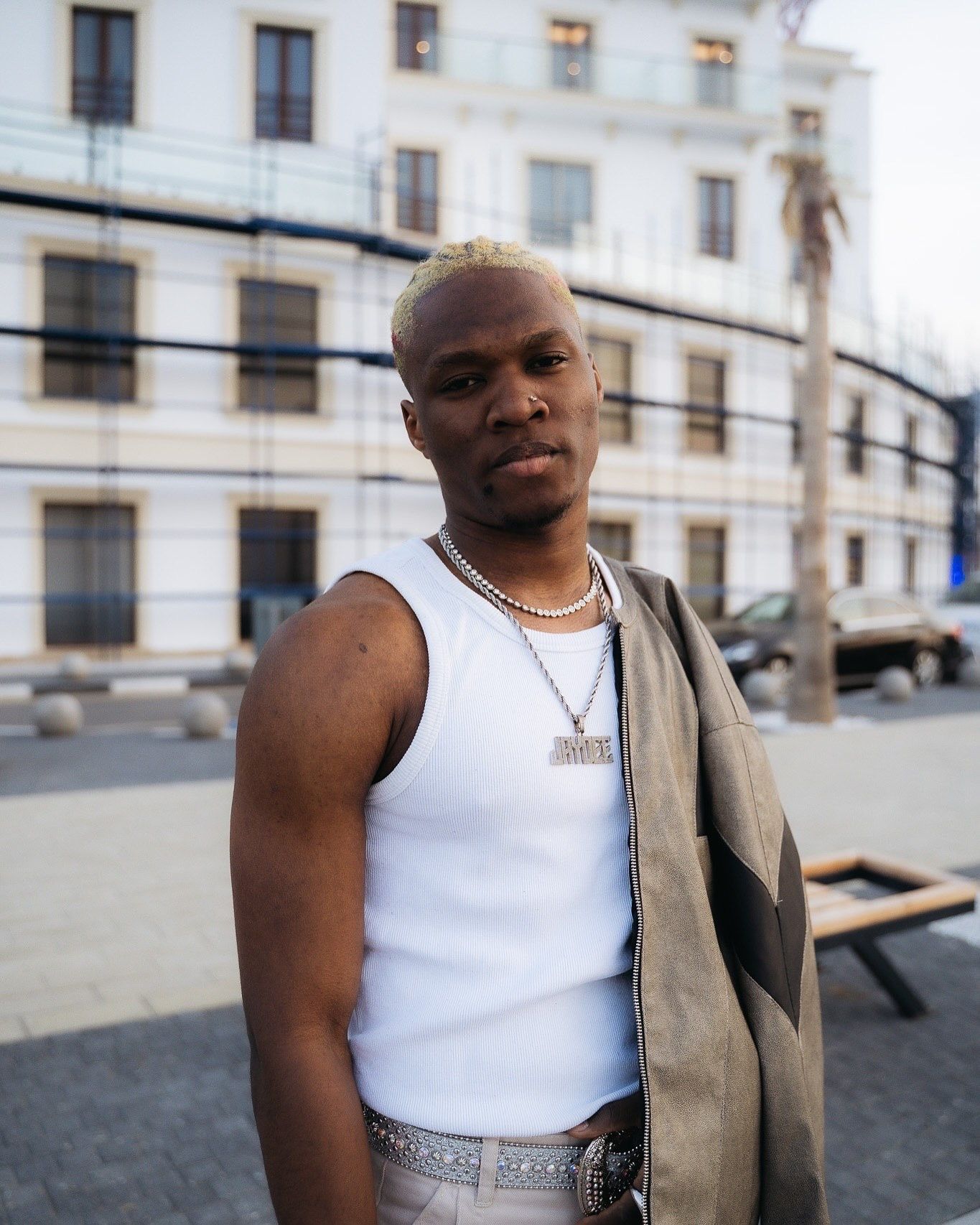 Young Black man with blonde hair wearing white tank top, pearl necklace, and chain pendant poses outdoors.