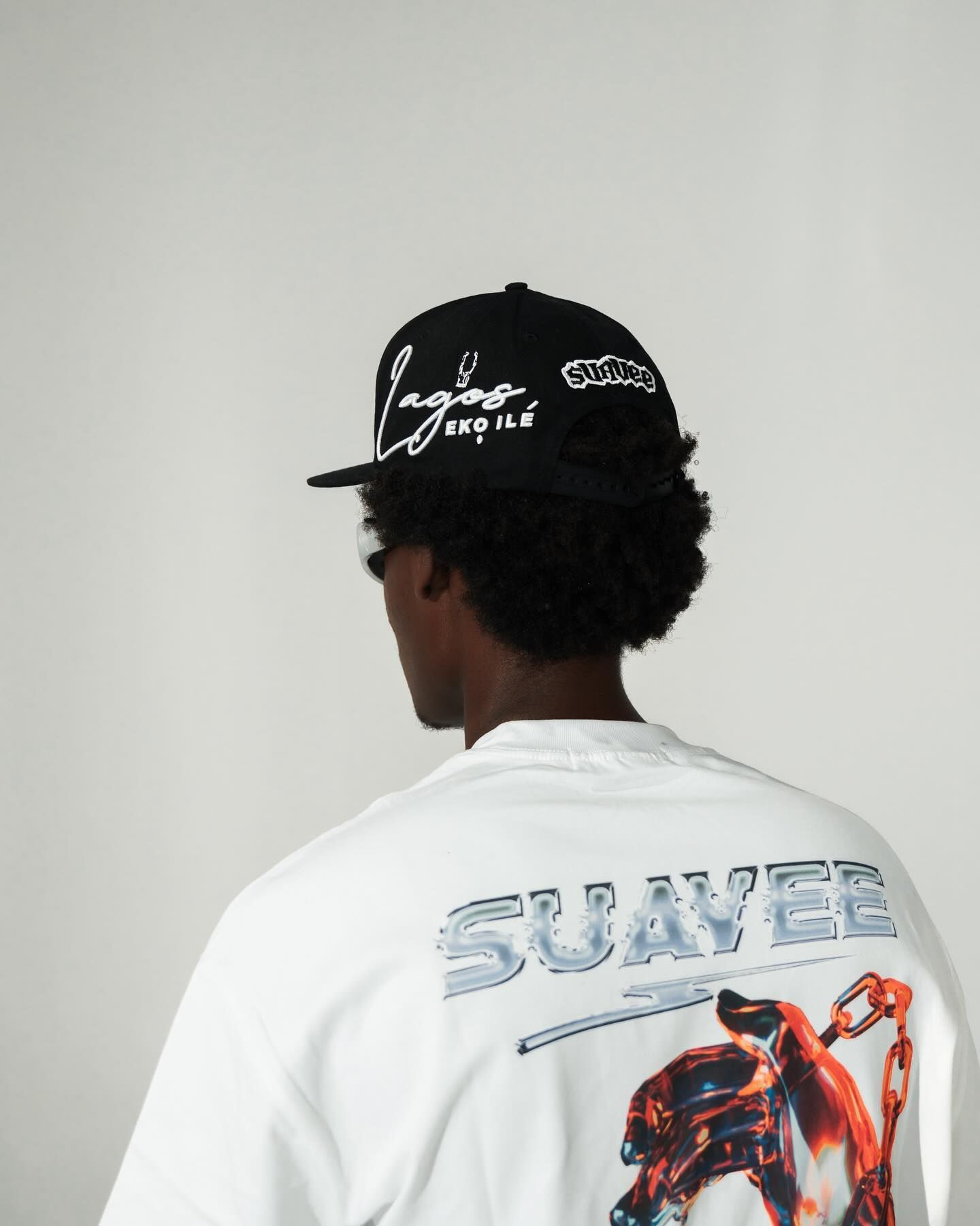 Man wearing black Lagos snapback cap and white Suavee graphic t-shirt, shown from behind against light background.