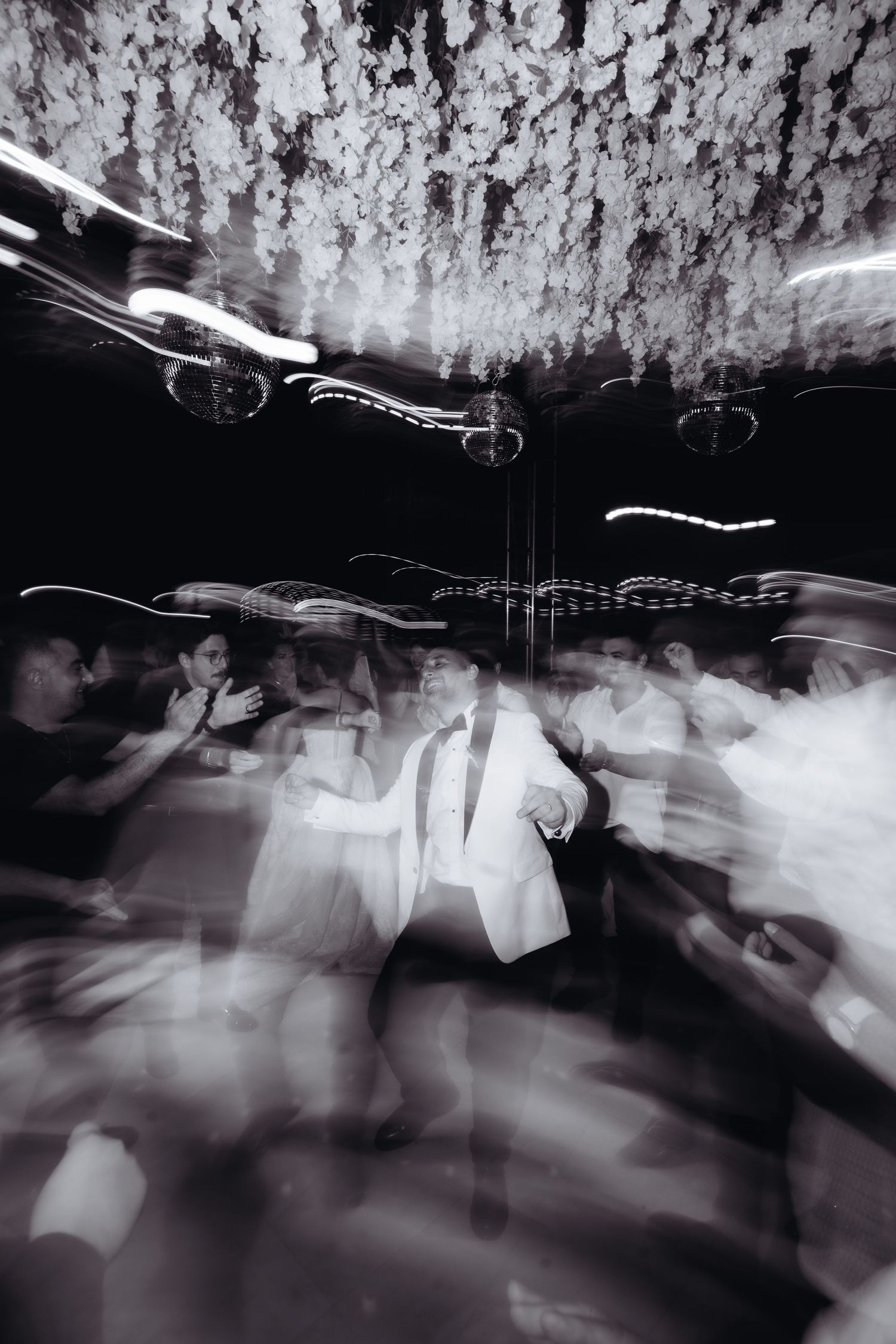 Black and white wedding reception dance floor photo with motion blur, couple in white attire dancing under floral ceiling.