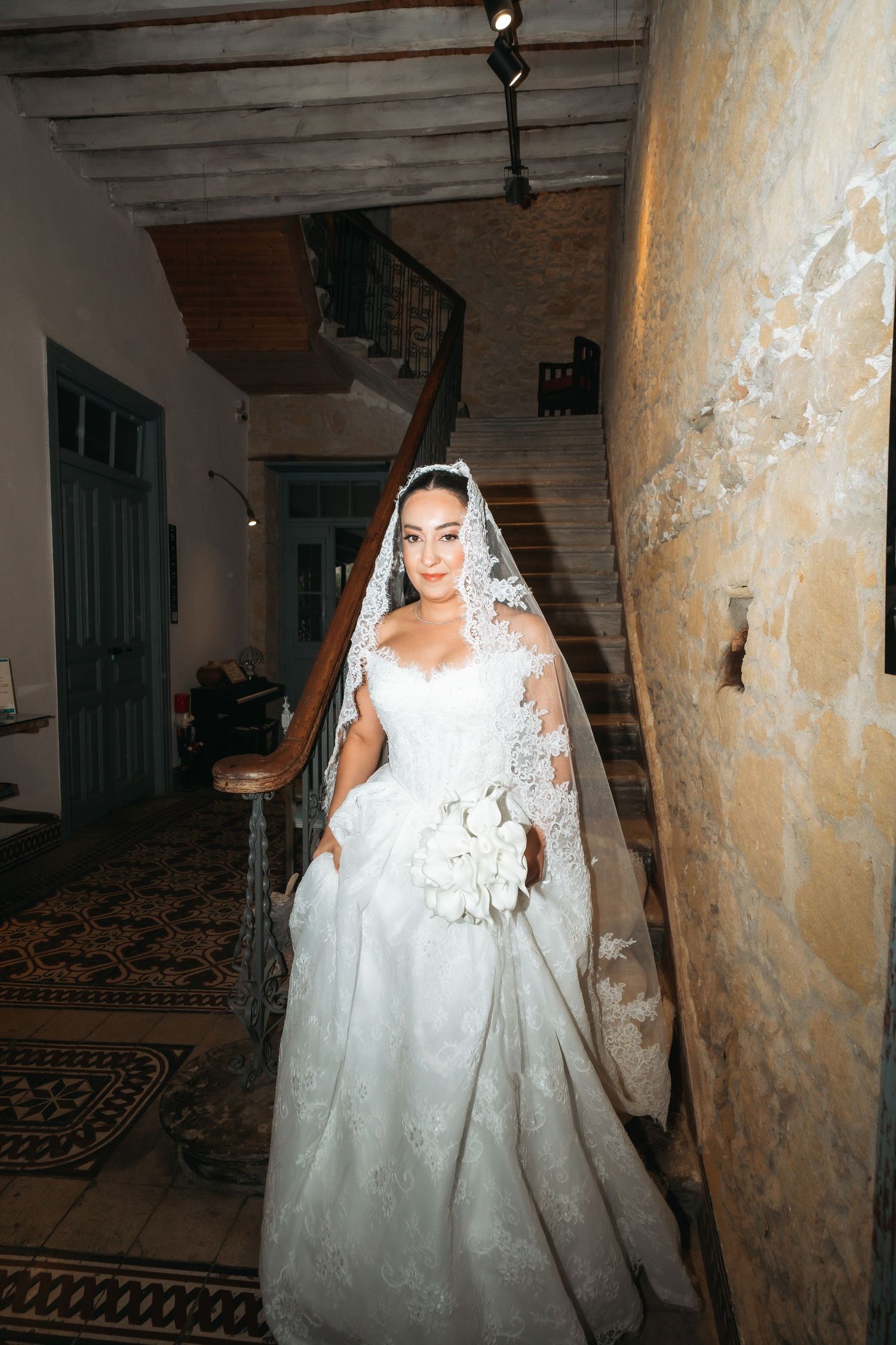 Bride in white lace gown and veil holds bouquet on staircase in elegant stone venue.