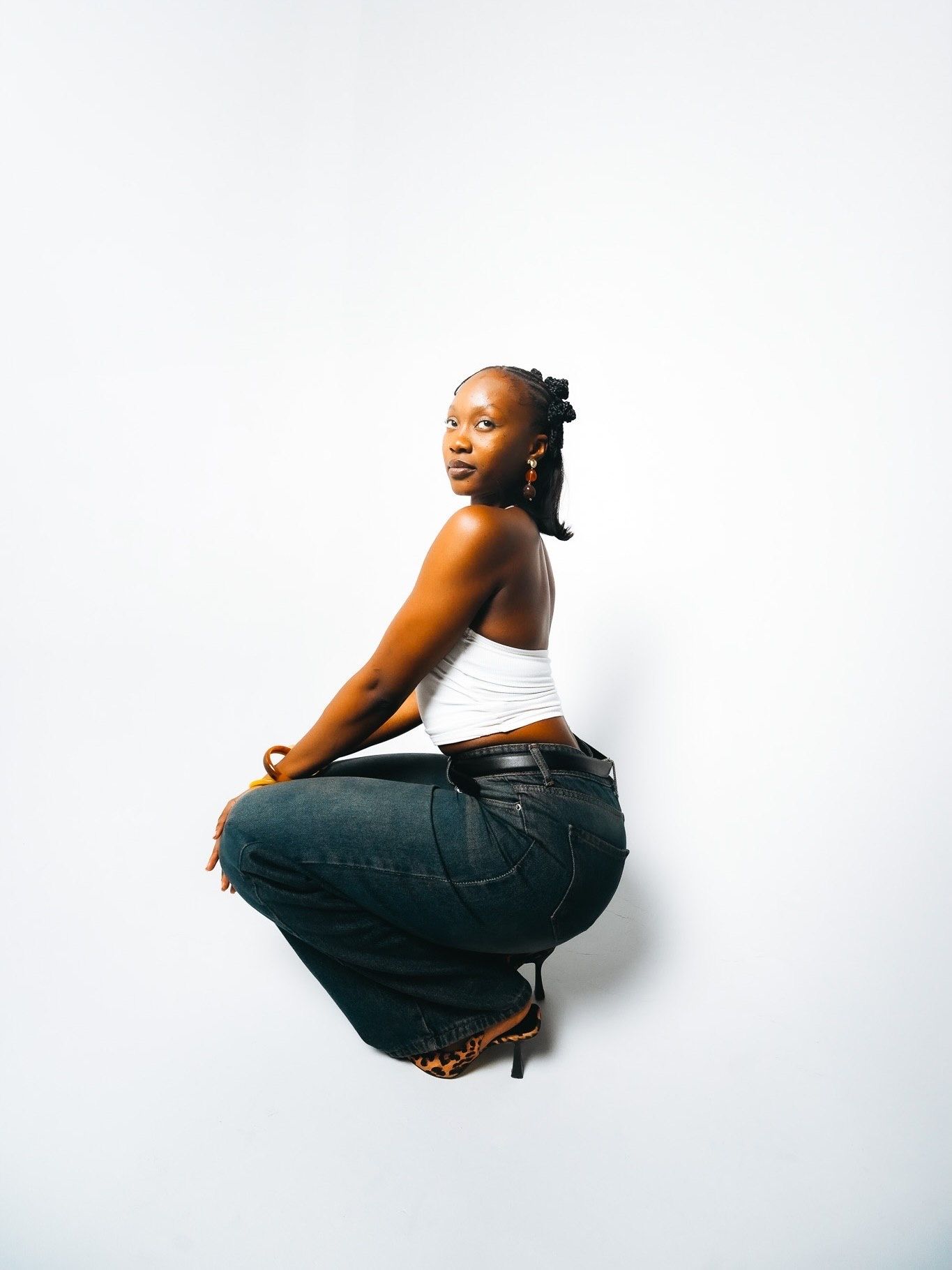 Black woman in white crop top and jeans crouches gracefully against white background in black and white photo.