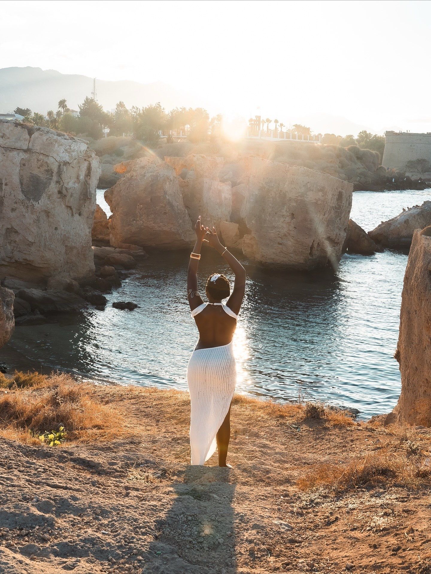 Woman in white dress raises arms toward golden sunset over rocky Mediterranean cove with calm blue waters.