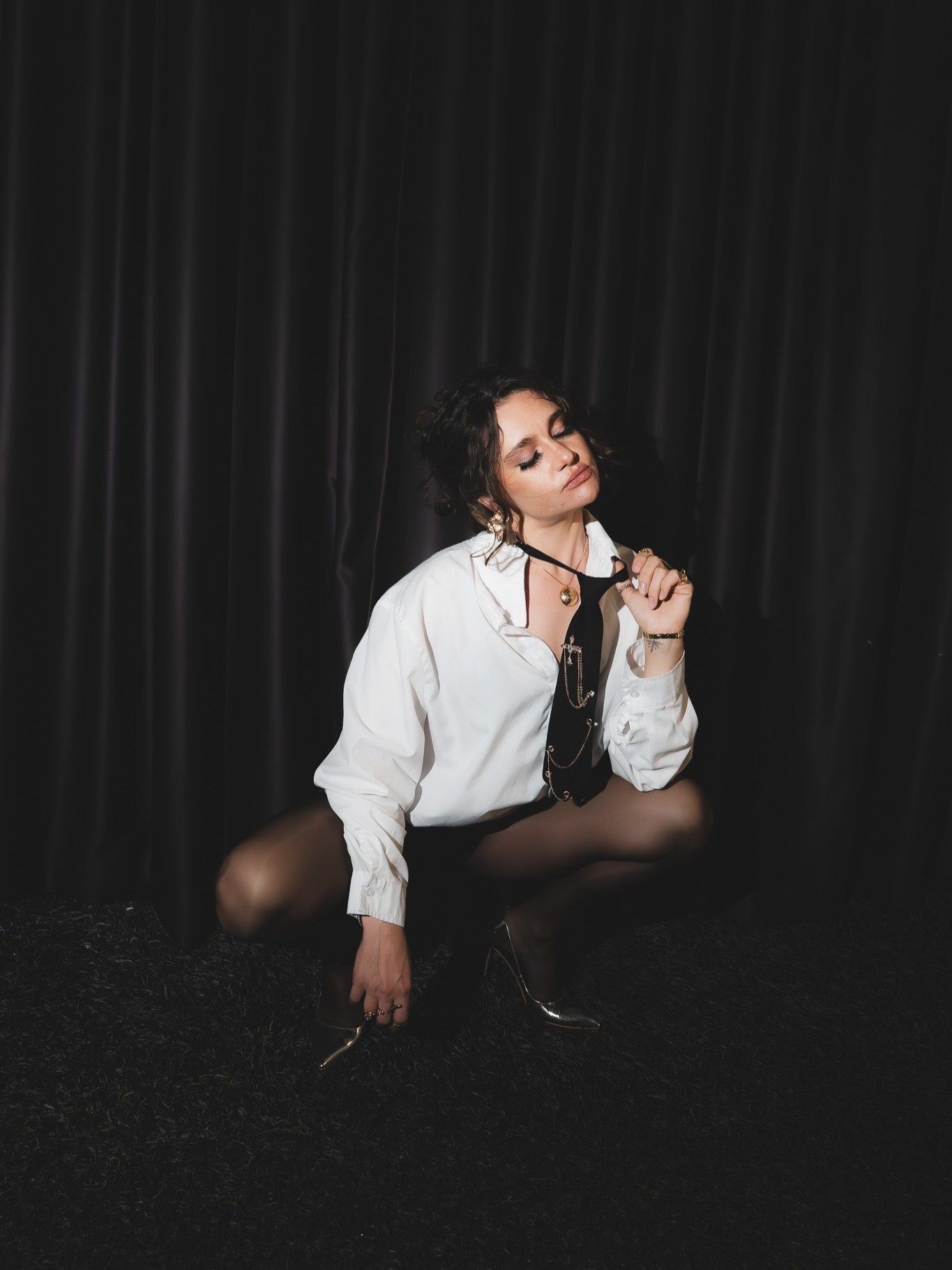 Young woman in white shirt and black tie poses seated on dark floor in moody editorial shoot.