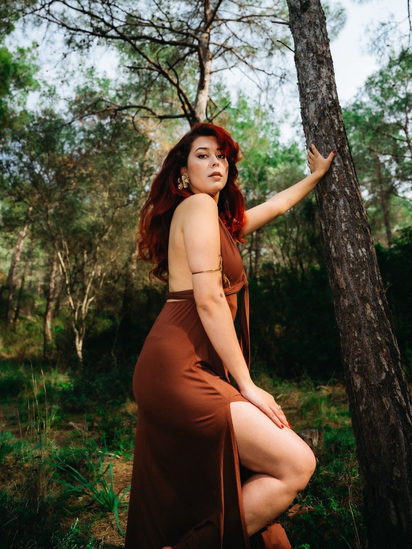 A woman in a brown dress poses gracefully against a tree in a lush forest setting.