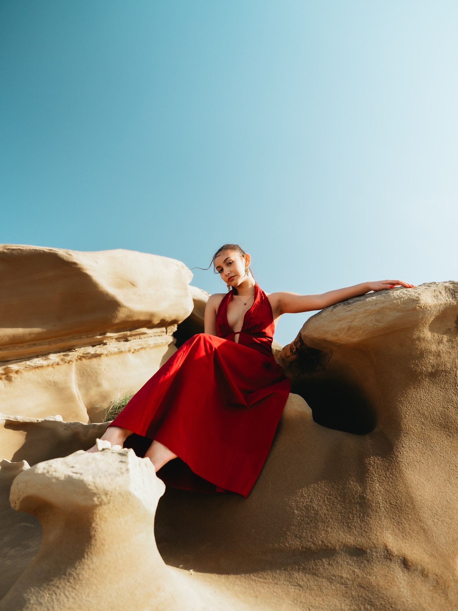 Woman in red dress poses gracefully on sandstone rocks under clear blue sky.