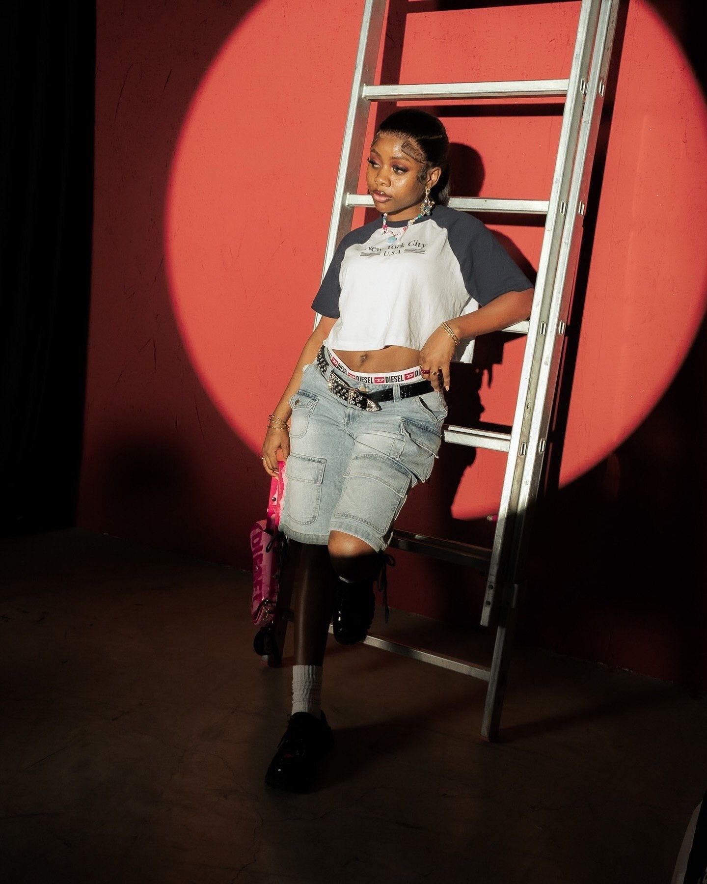 Young woman in crop top and denim shorts leaning against a ladder in a red spotlight.