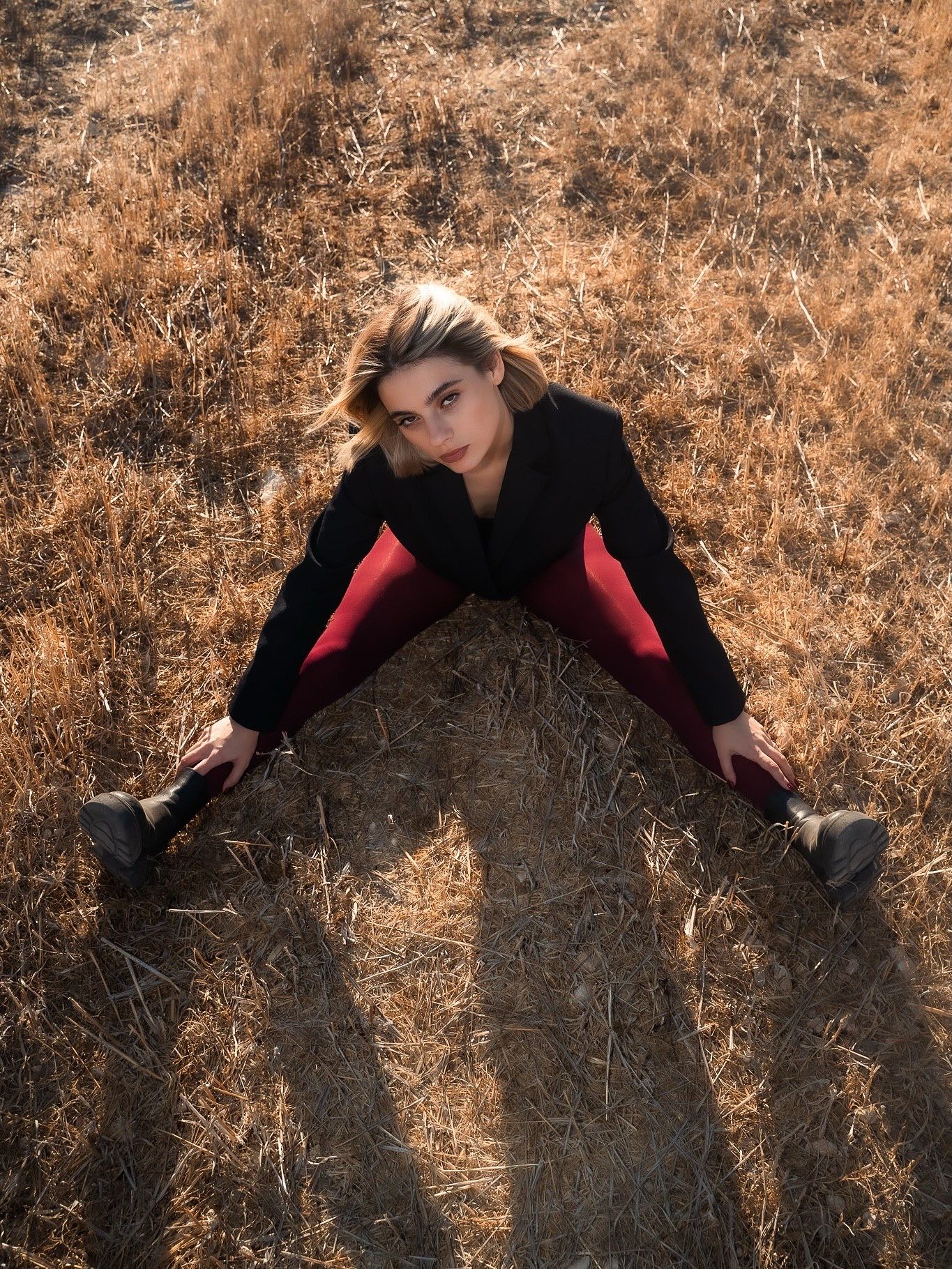Woman in black jacket and red pants doing splits in a dry golden grass field, aerial view.