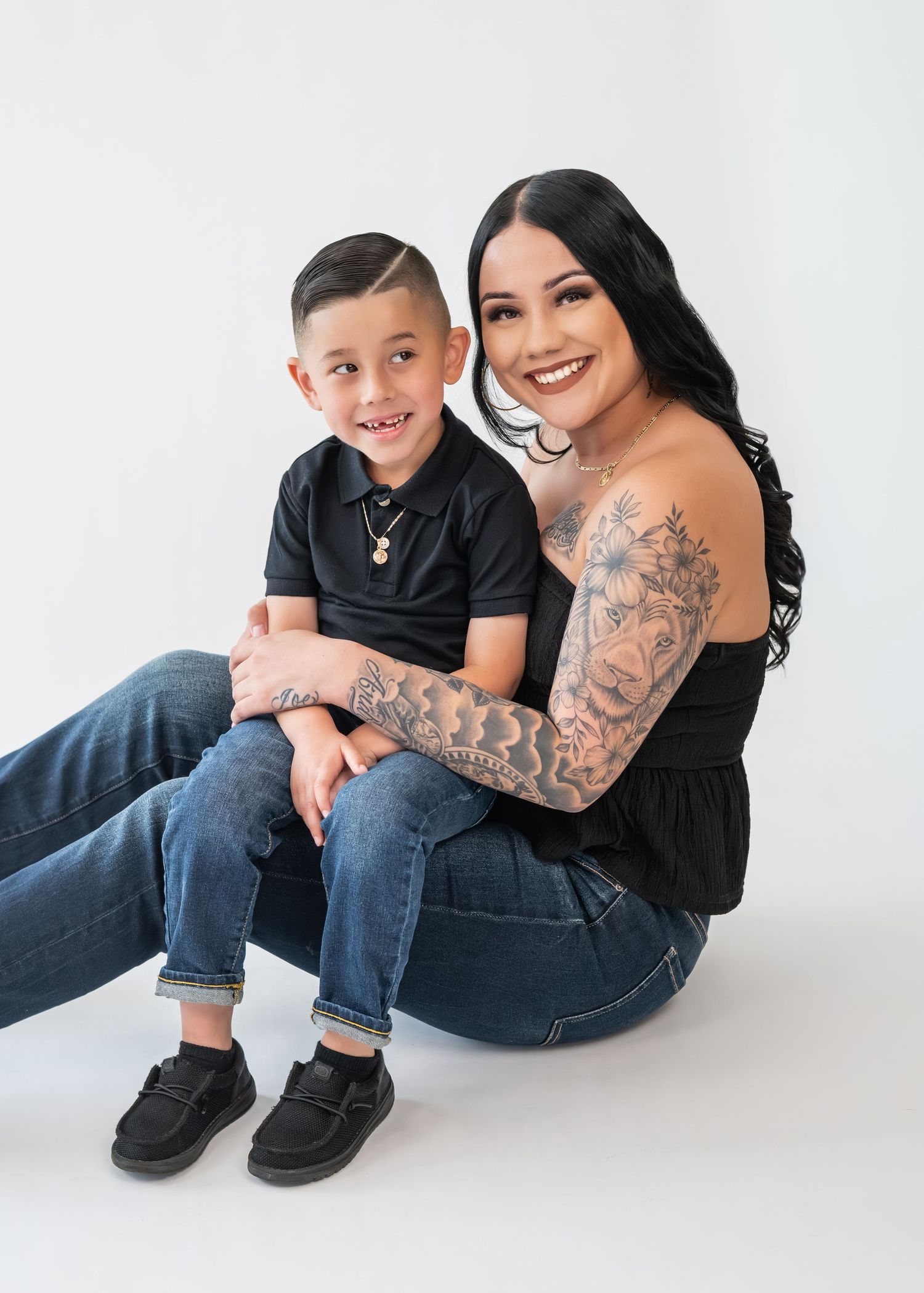 A tattooed woman smiles while holding a young boy in a black polo shirt, both wearing jeans, posed against a white background.