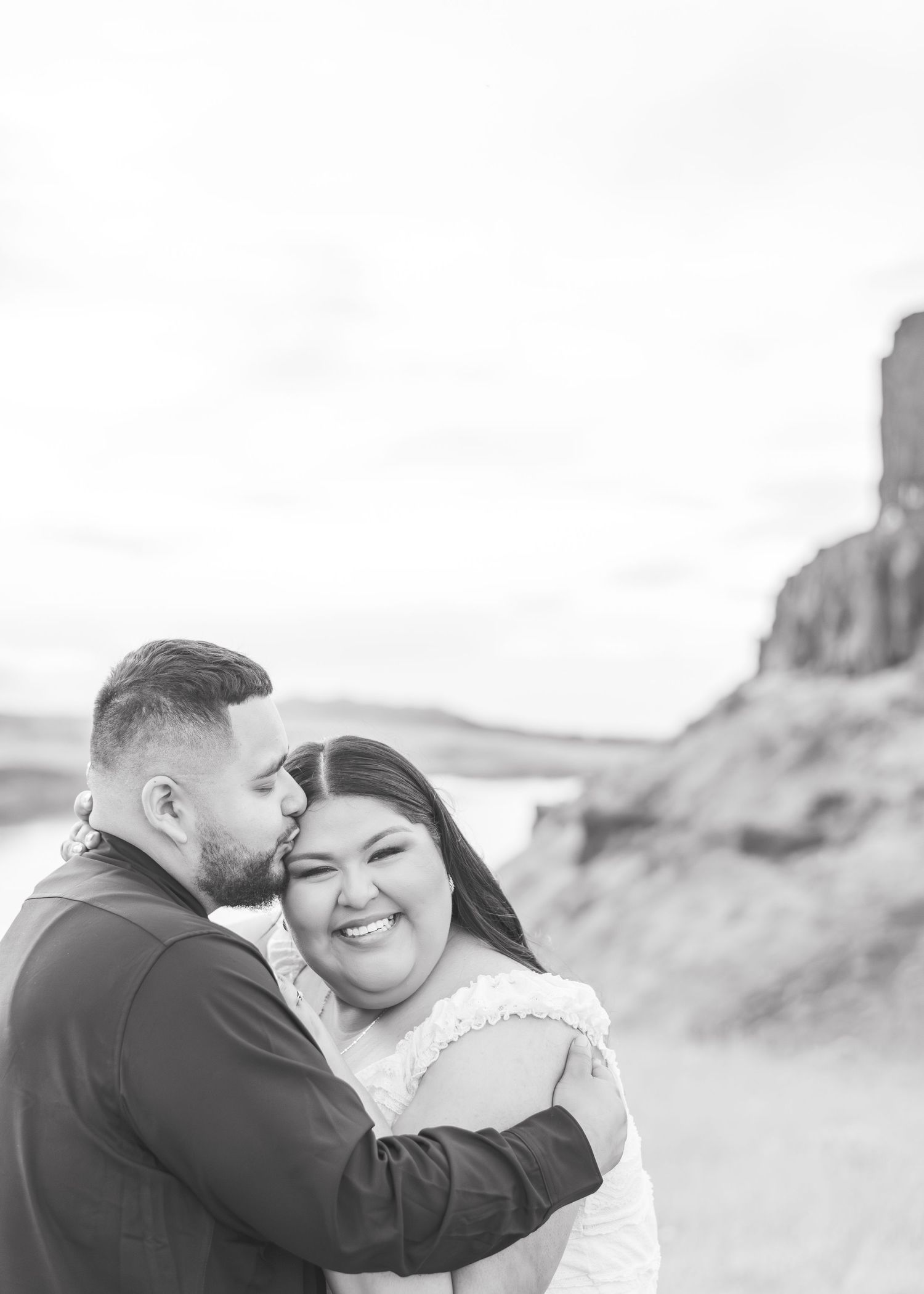 Black and white photo of a smiling bride being kissed on the cheek by groom outdoors near a rocky landscape.
