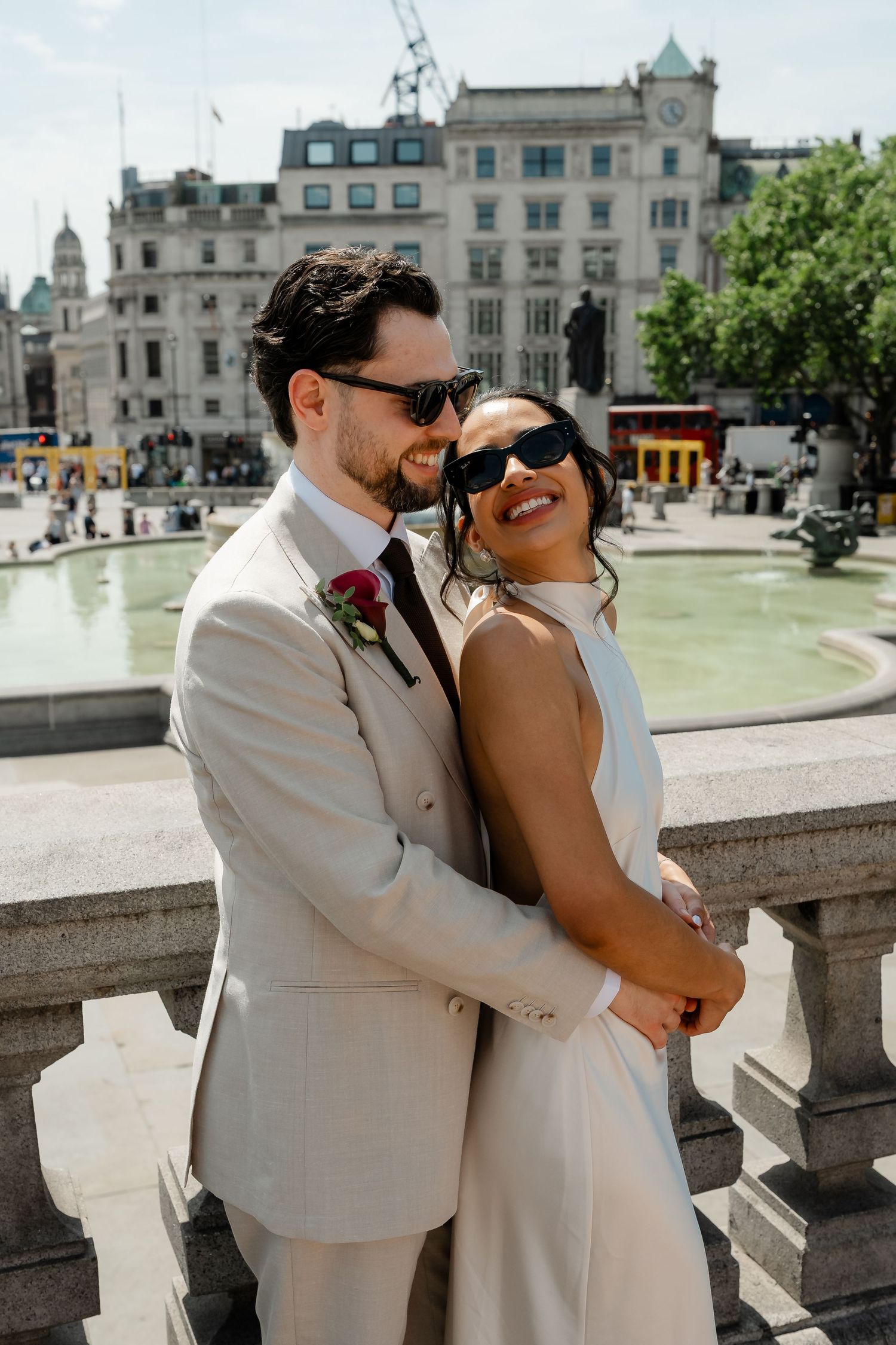 Smiling newlyweds in sunglasses embrace near Trafalgar Square fountain in London on their wedding day.