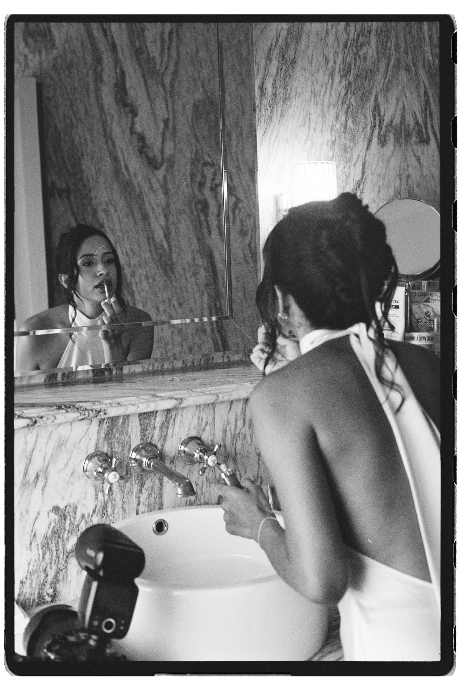 Black and white film photo of bride applying lipstick at a marble vanity mirror before her wedding ceremony.